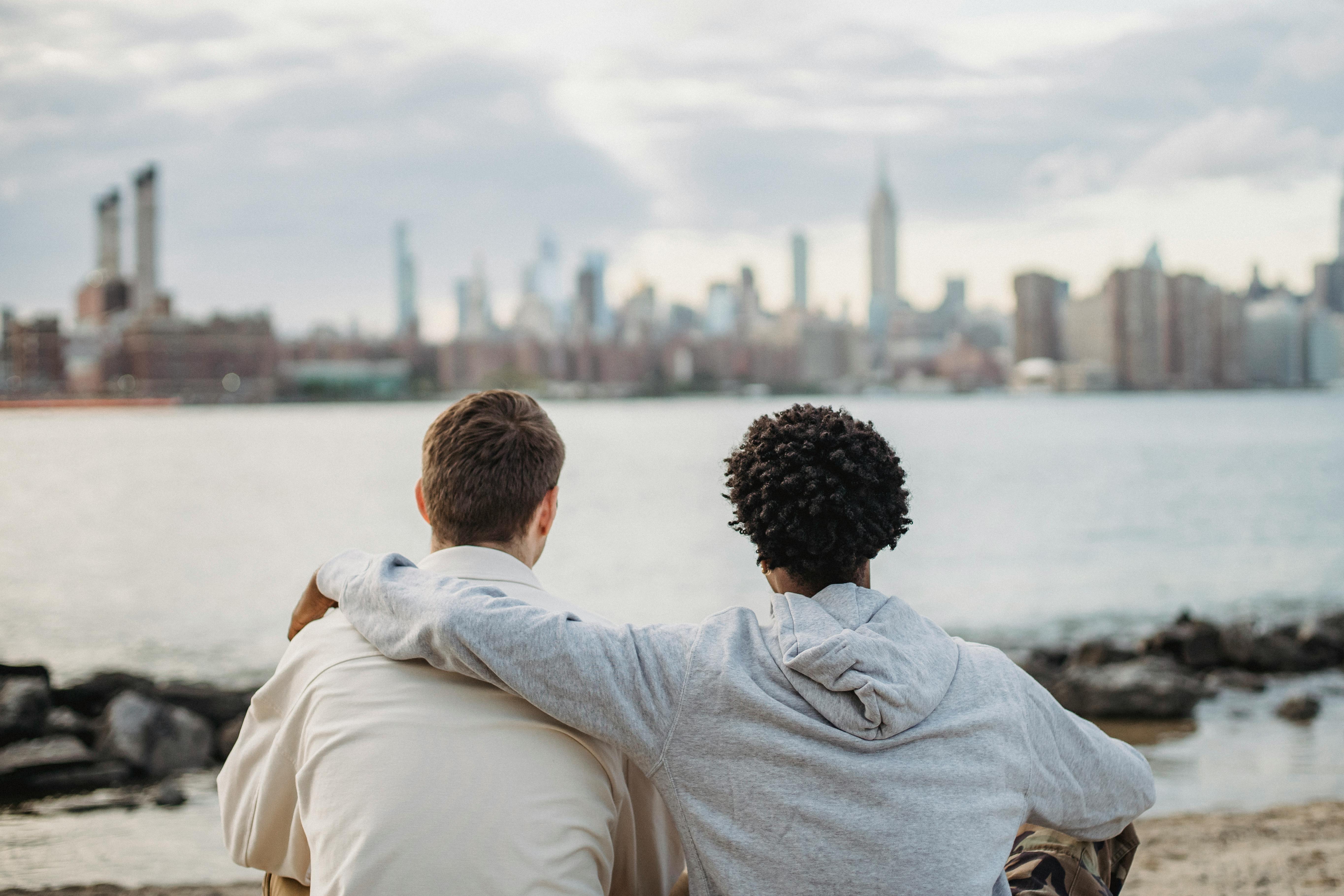 Zwei Personen sitzen am Ufer und blicken auf die Skyline einer Stadt mit Wolkenkratzern im Hintergrund.