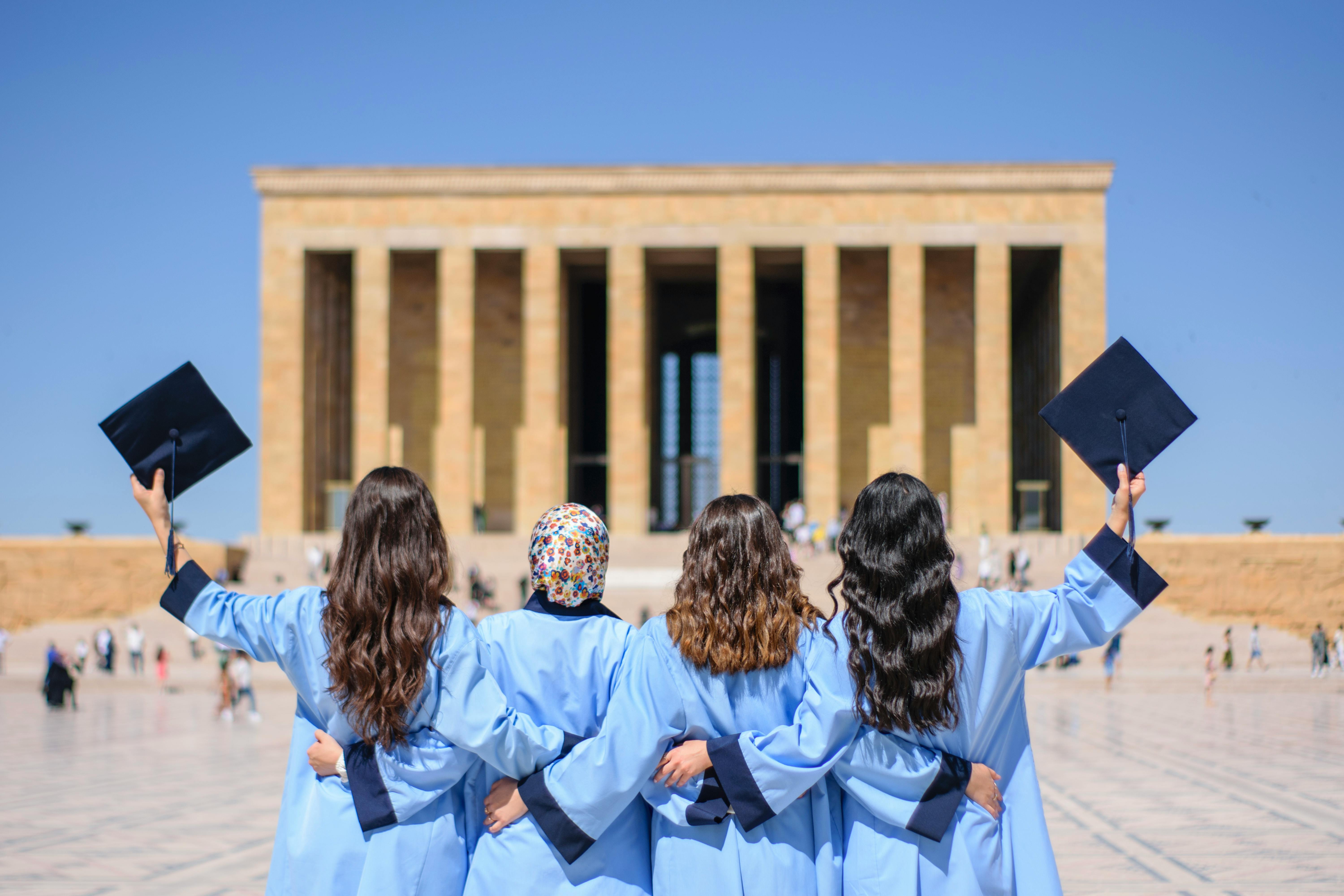 Vier Absolventinnen in blauen Roben und Hüten stehen mit dem Rücken zur Kamera vor einem monumentalen Gebäude.