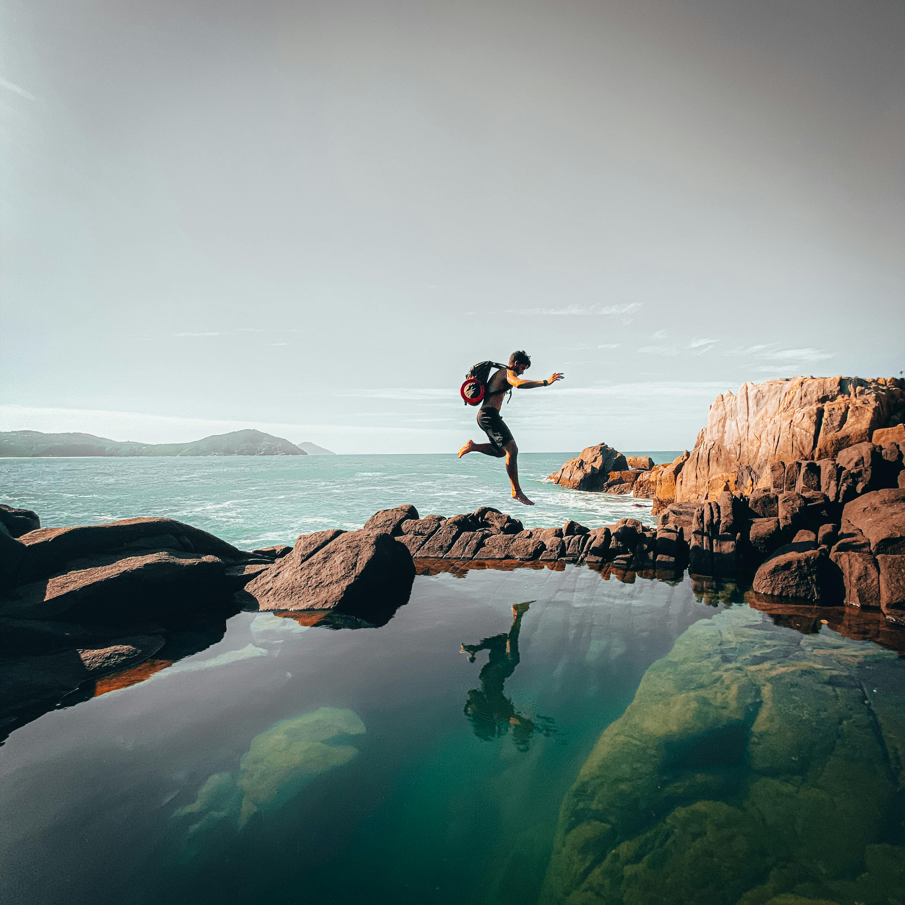 Eine Person springt von einem Felsen ins Wasser, umgeben von einer Küstenlandschaft mit Bergen im Hintergrund.