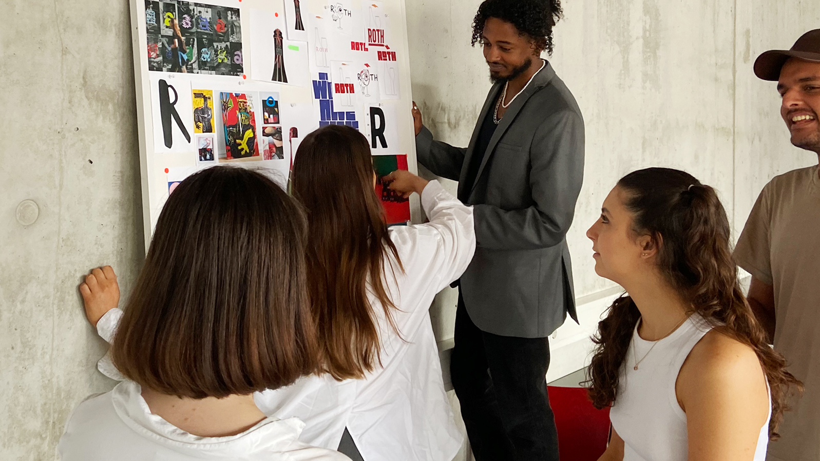 A group of five people stands around a large poster displaying various images and texts. One person points at the poster while the others watch attentively. The atmosphere feels interactive and collaborative.
