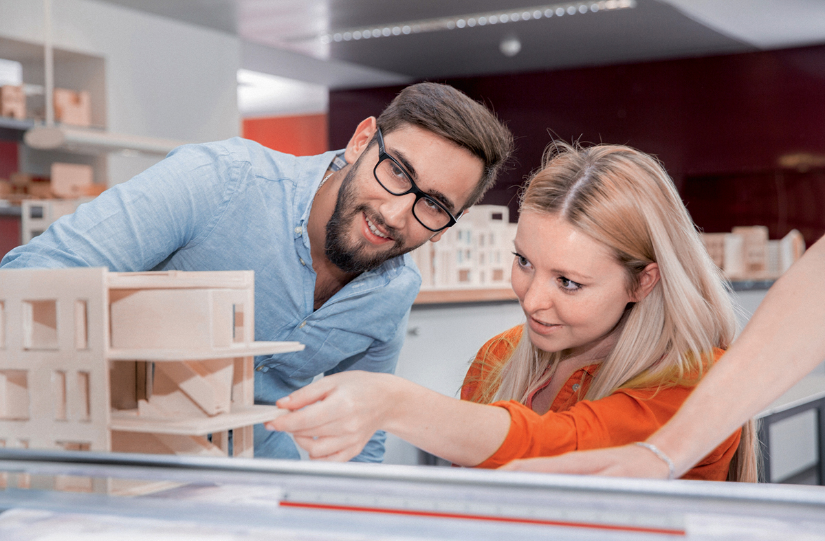 Zwei Personen betrachten ein Architekturmodell. Der Mann zeigt auf einen Teil des Modells, während die Frau aufmerksam zuschaut. Beide scheinen in einem kreativen Arbeitsumfeld zu sein, umgeben von weiteren Modellen und Materialien.