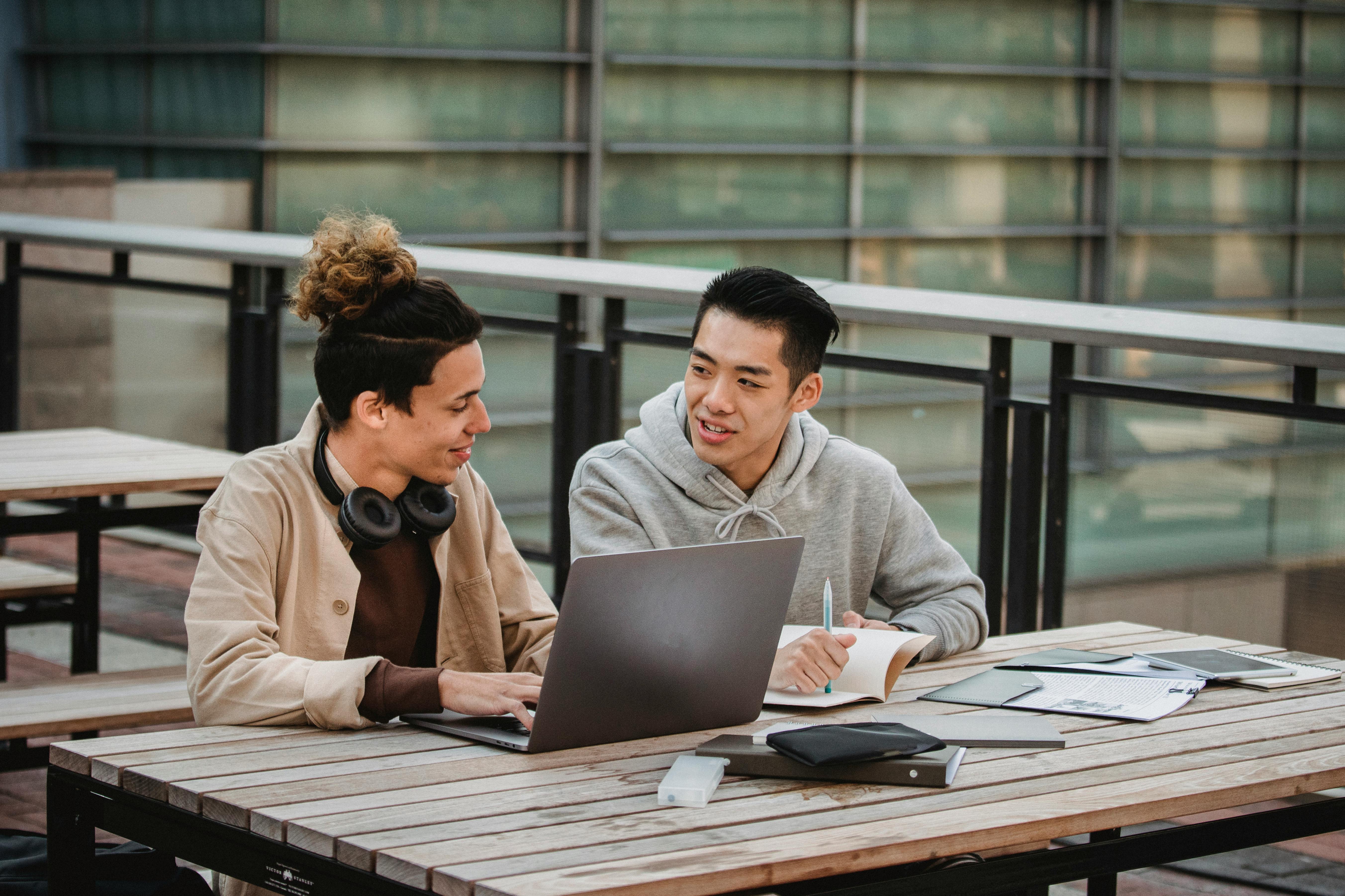 Two young men are sitting at an outdoor table, working together on a laptop. The man on the left is wearing headphones and has a relaxed expression, while the man on the right is taking notes and looking at the screen.