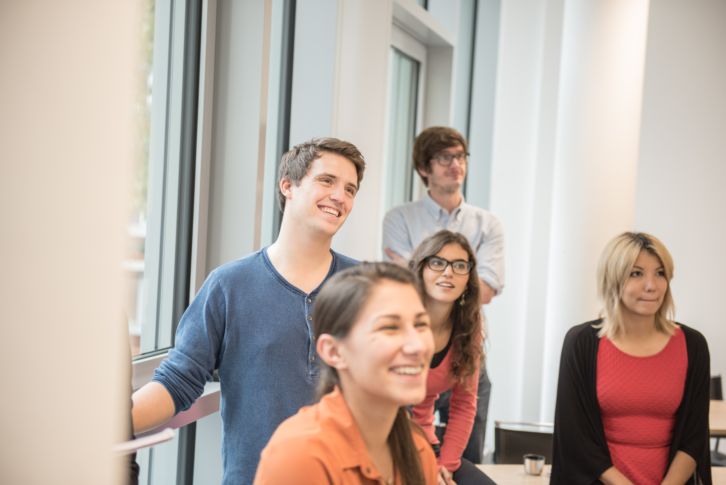 A group of five young adults stands in a bright room. They are smiling and looking attentively in one direction. Two of them are wearing glasses, while the others are dressed casually.