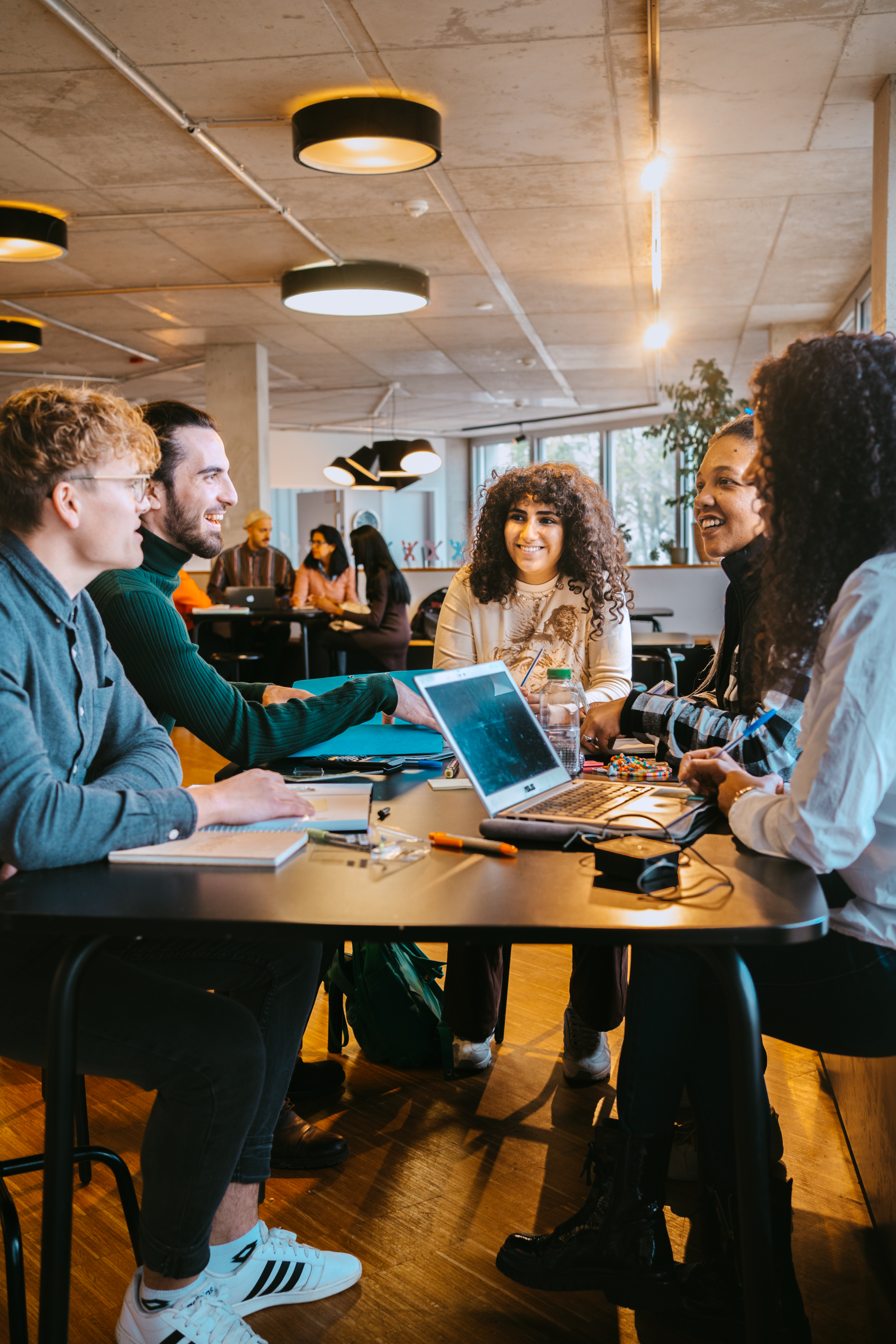 A group of five people is sitting at a table, surrounded by laptops and notebooks. They are discussing and collaborating in a modern, bright room with large windows and a relaxed atmosphere.