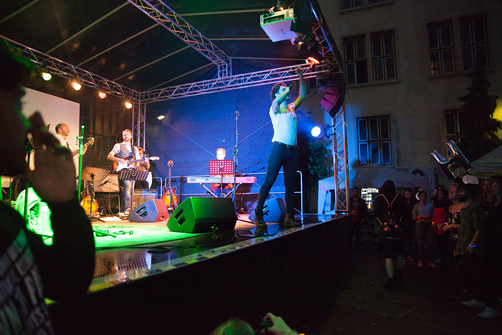 A musician performs on stage, surrounded by instruments and band members. The audience stands in front of the stage, watching the performance. Colorful lights illuminate the scene.