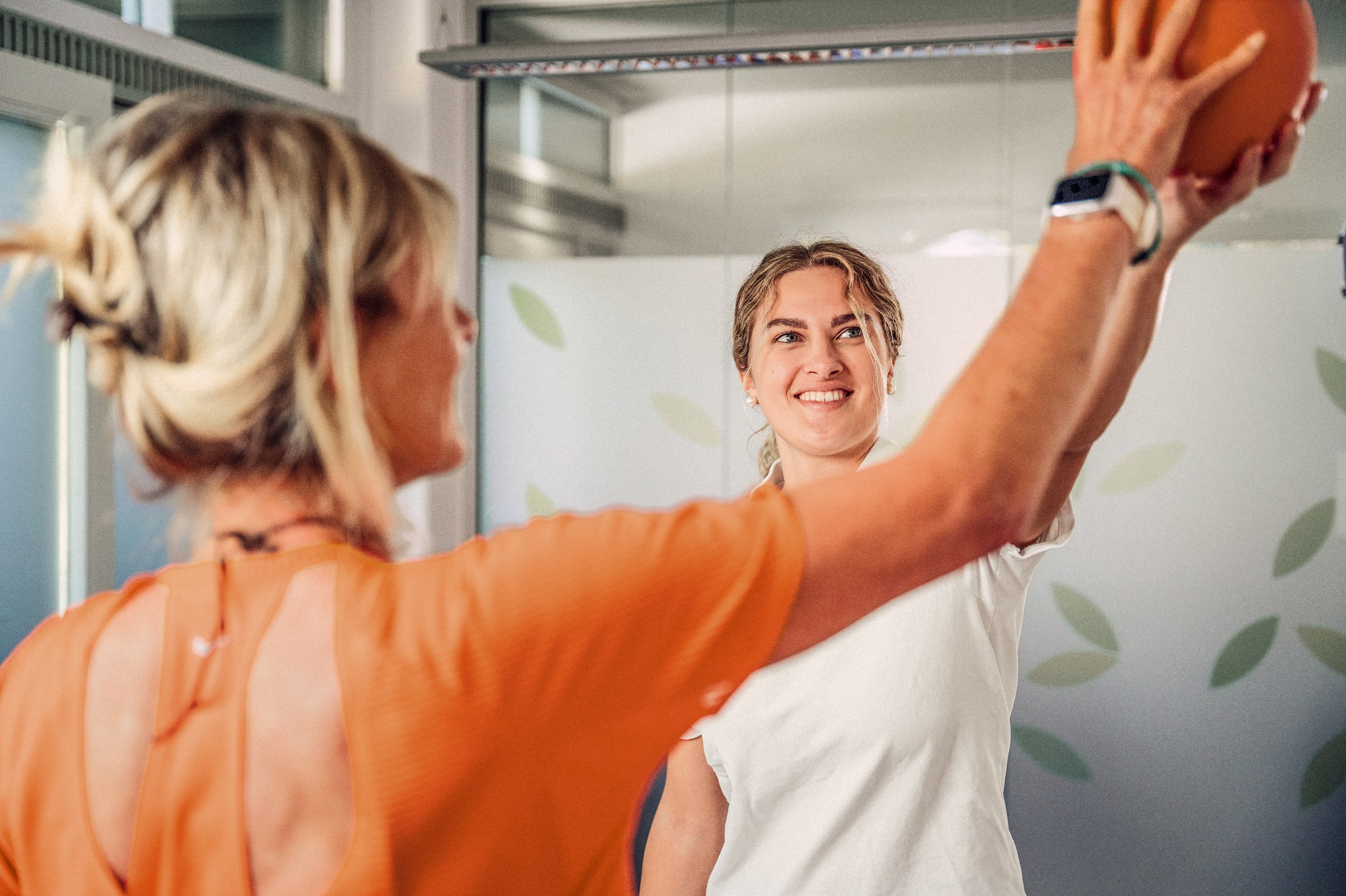 A person is holding a ball up while another person watches in a training room. Both are wearing athletic clothing. The room has a bright, cheerful atmosphere with plant motifs in the background.