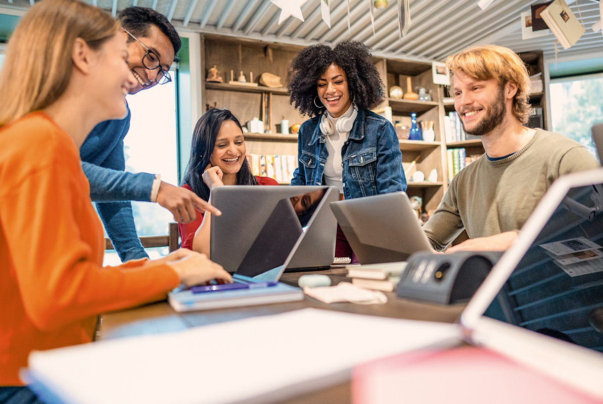 A group of five people is sitting at a table, looking at laptops. They seem to be working on a project and discussing with each other. The atmosphere feels collaborative and friendly.