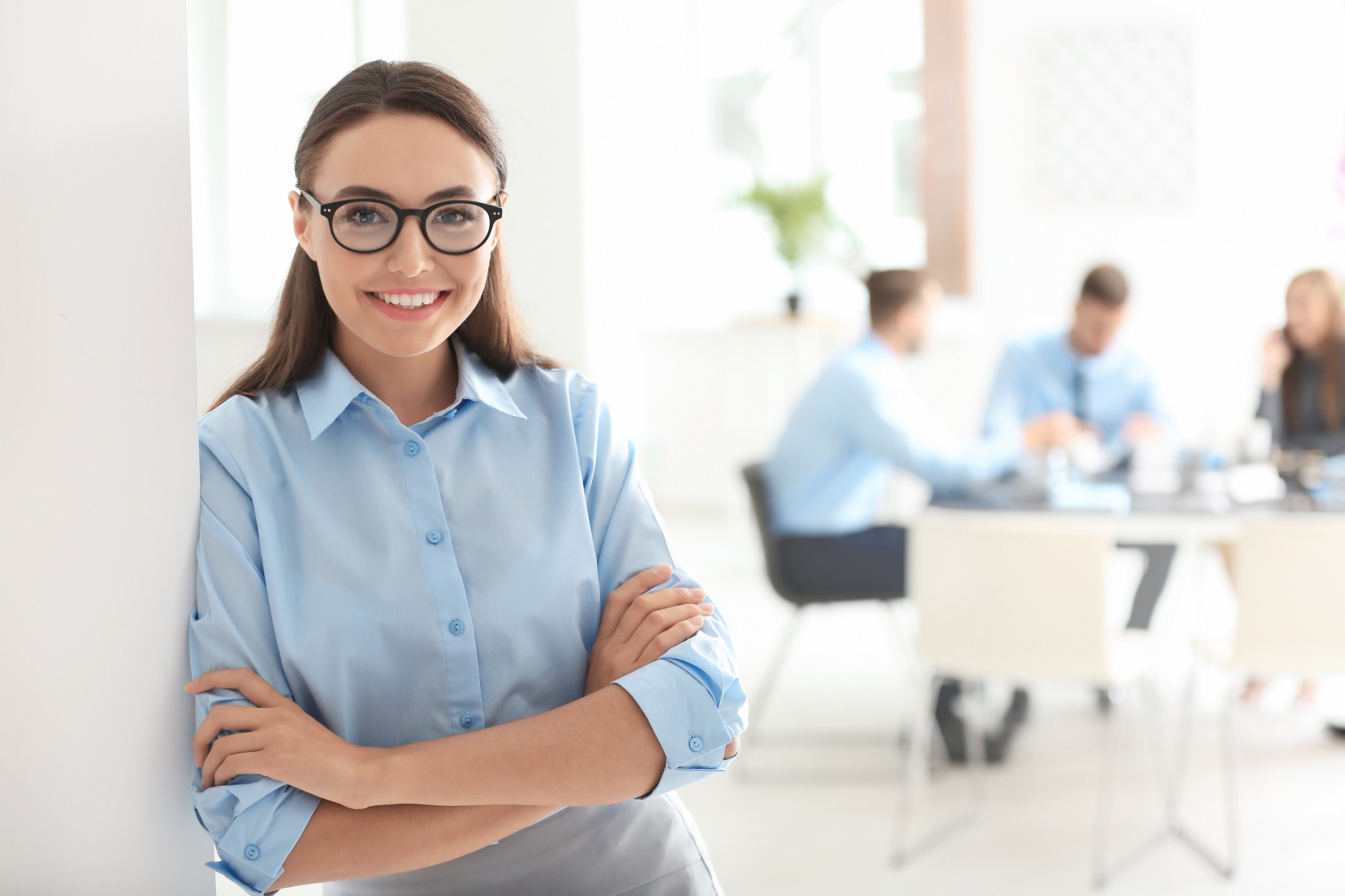Eine Frau mit Brille und blauer Bluse steht lächelnd an einer Wand. Im Hintergrund sind mehrere Personen in einem Besprechungsraum zu sehen, die an einem Tisch sitzen und miteinander sprechen.