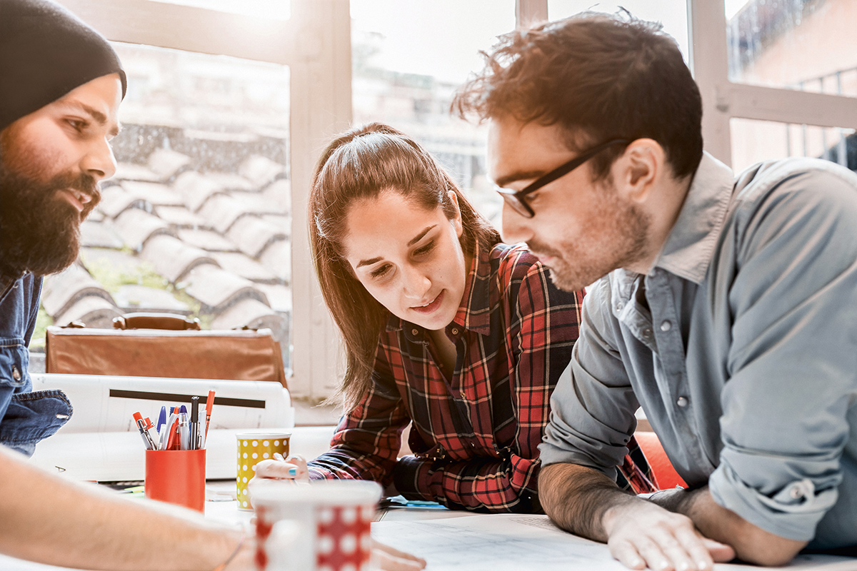 Three people are working together at a table, taking notes and exchanging ideas. One person is wearing a hat, another has on a checkered shirt, and the third is wearing glasses.