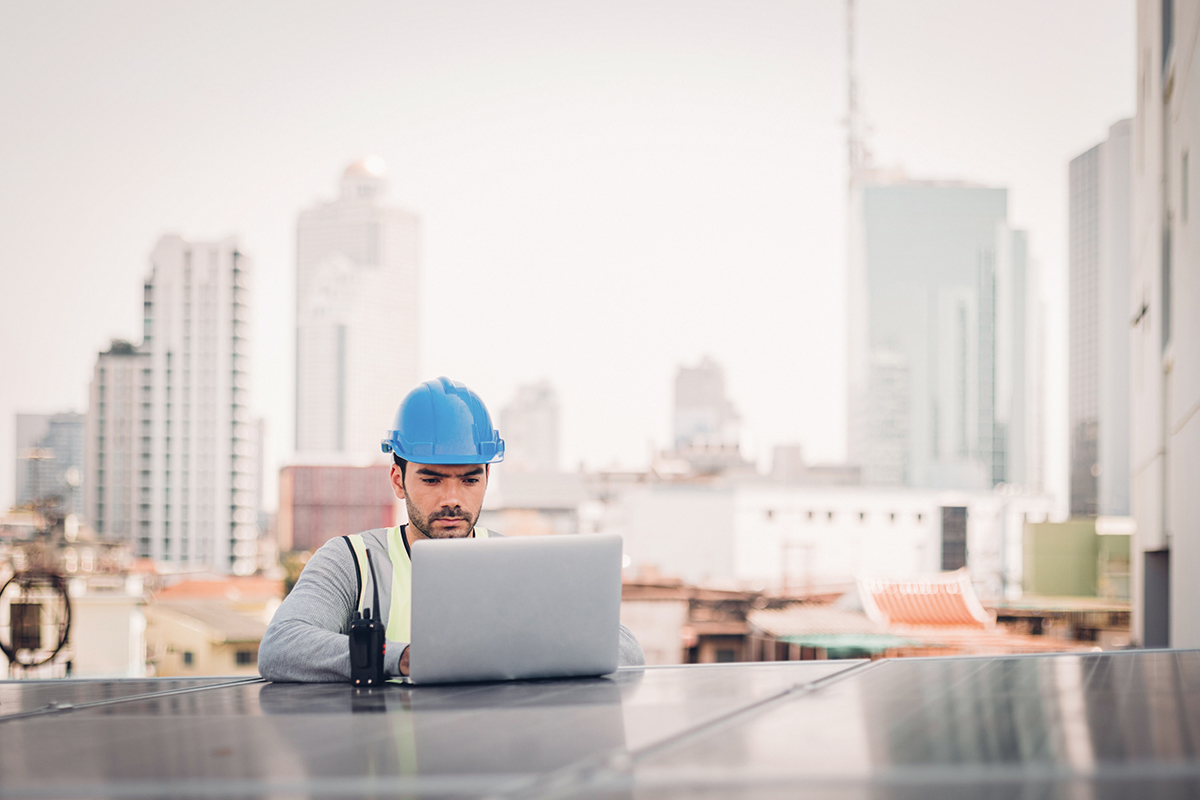A construction worker wearing a blue helmet and a safety vest is working on a laptop on a rooftop. In the background, modern buildings and an urban landscape can be seen. A walkie-talkie is lying next to him.
