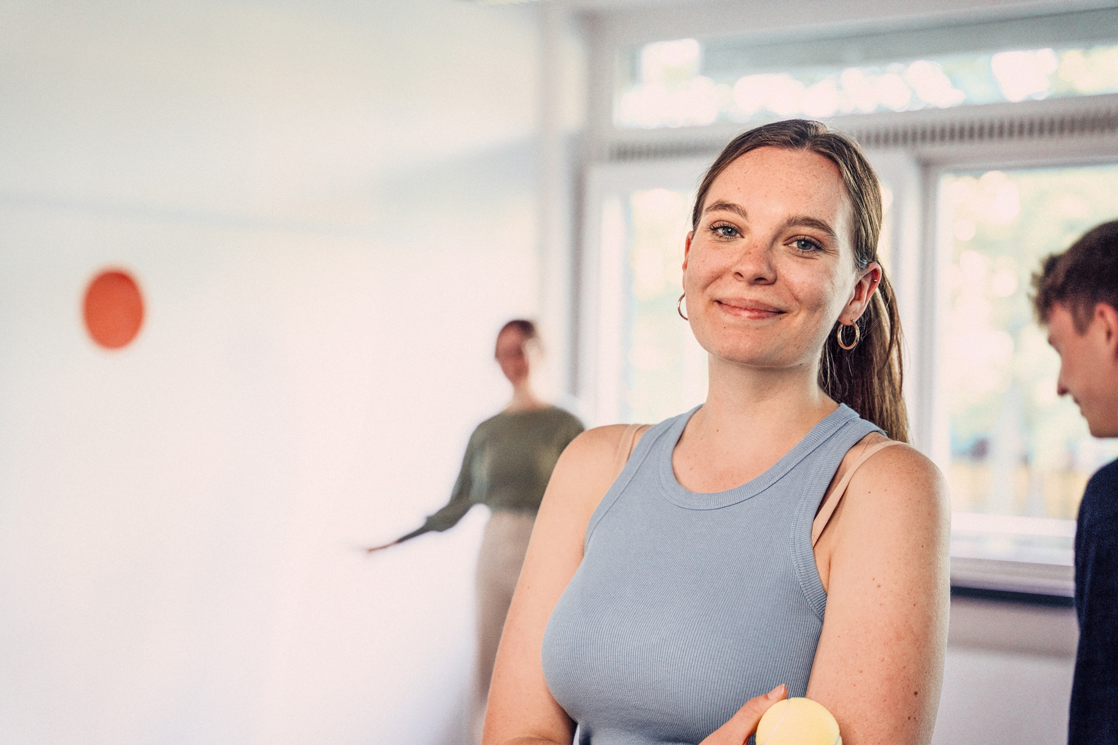 A woman with long hair and a blue top smiles at the camera while holding a yellow ball. In the background, two people can be seen chatting in a bright room.