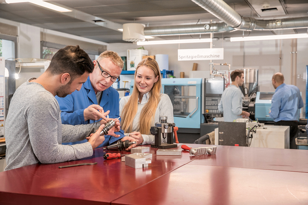 Two people are examining a technical device while a third man gives them instructions. In the background, machines and tools are visible, indicating a technical environment.
