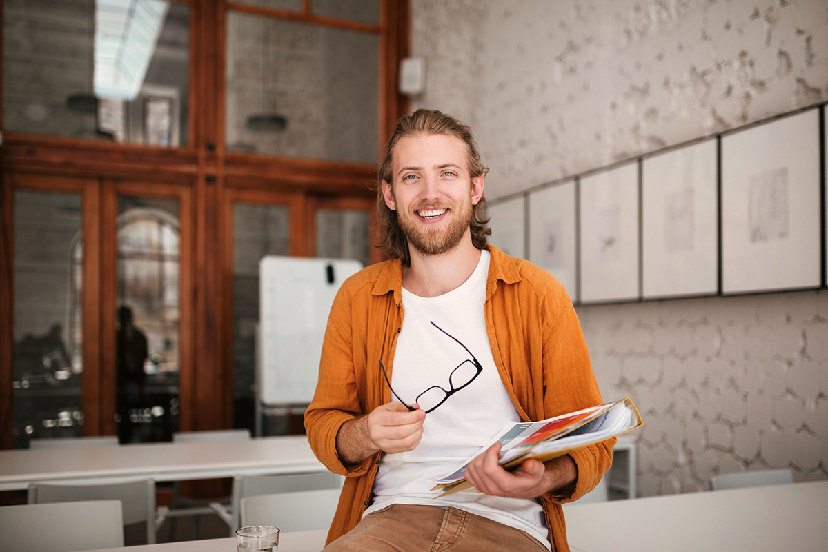 A young man with long hair and a beard is sitting on a table in a modern room. He is holding a folder with documents and a pair of glasses in his hand. He is wearing an orange shirt and a white T-shirt.