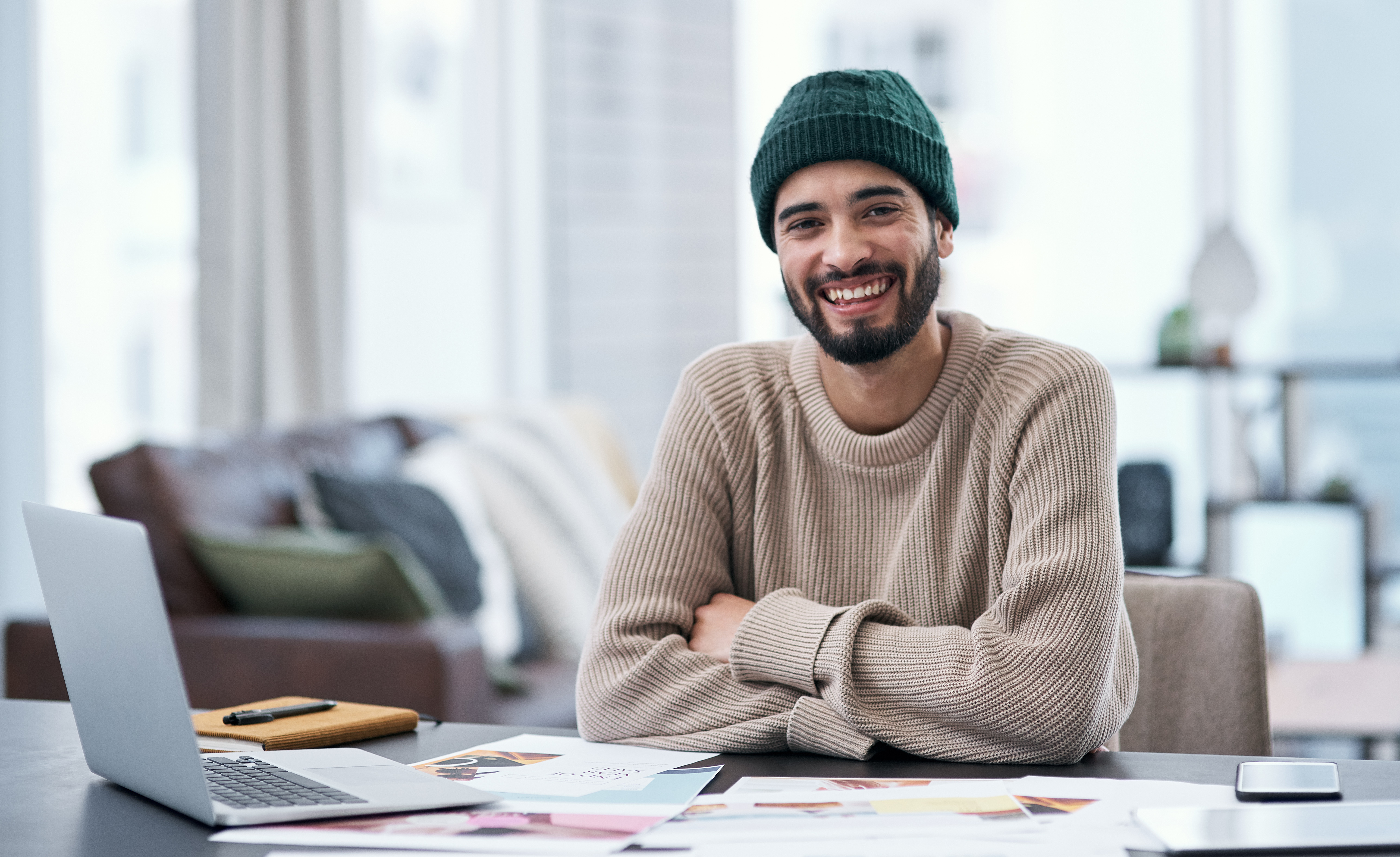 A smiling man with a beard is wearing a green hat and a beige sweater, sitting at a table with a laptop and some documents.