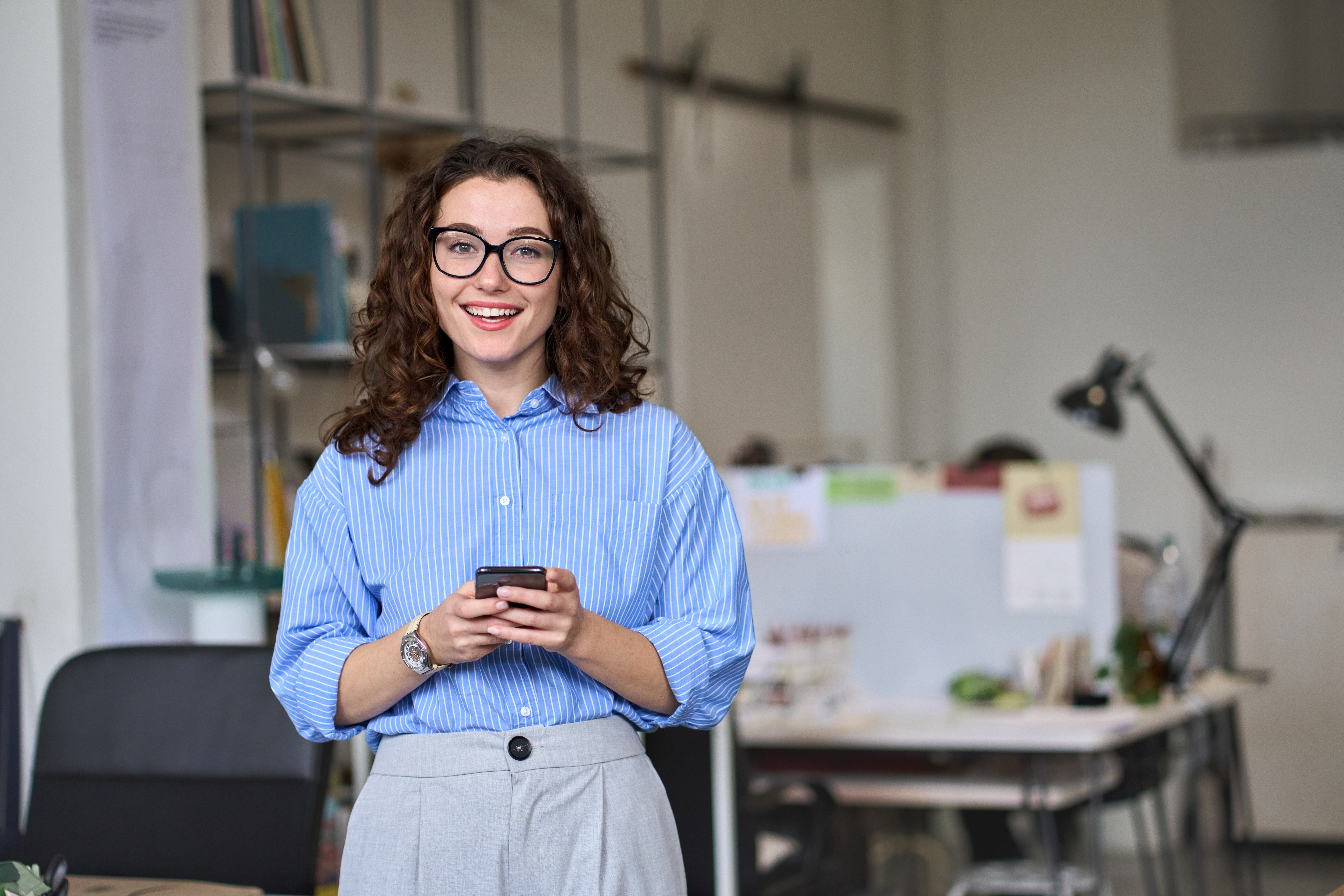 Eine Frau mit lockigem Haar und Brille steht in einem modernen Büro und hält ein Smartphone in der Hand. Sie trägt ein gestreiftes Hemd.