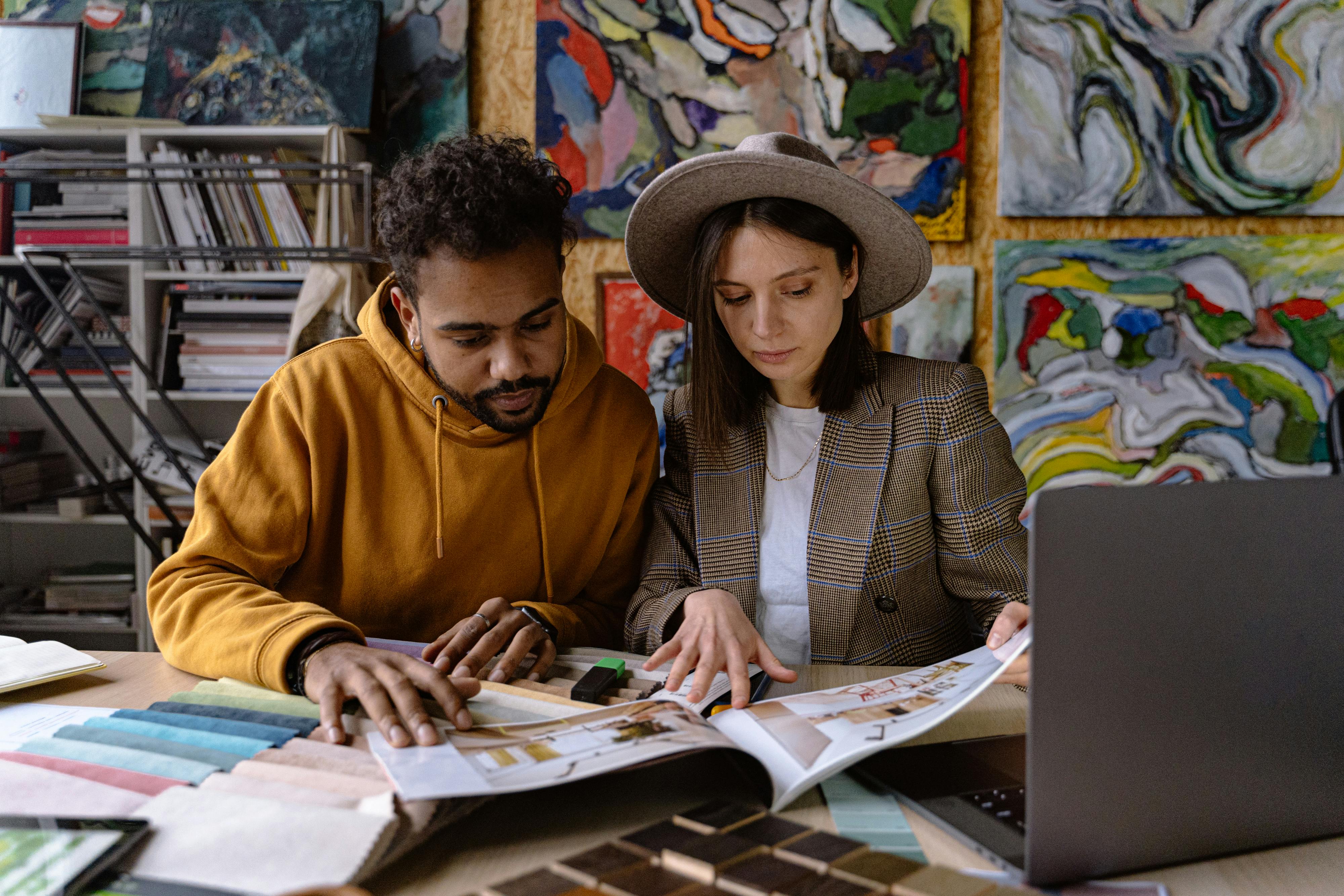 Ein Mann und eine Frau betrachten gemeinsam ein Magazin auf einem Tisch. Um sie herum liegen verschiedene Stoffmuster und Farbpaletten. Im Hintergrund sind bunte Gemälde an der Wand zu sehen. Ein Laptop steht ebenfalls auf dem Tisch.