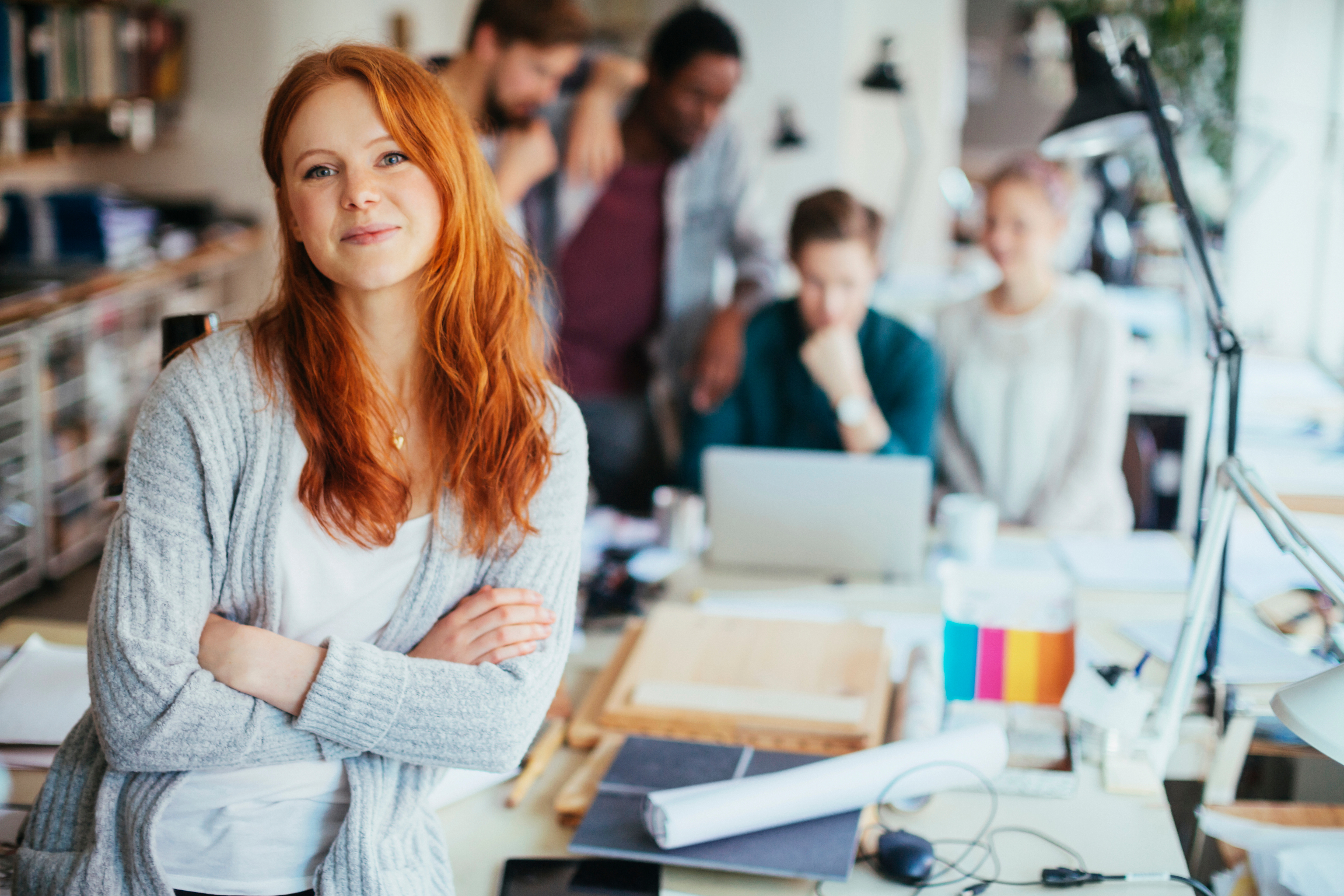 Eine lächelnde Frau mit roten Haaren steht im Vordergrund, während mehrere Personen im Hintergrund an einem Tisch arbeiten.