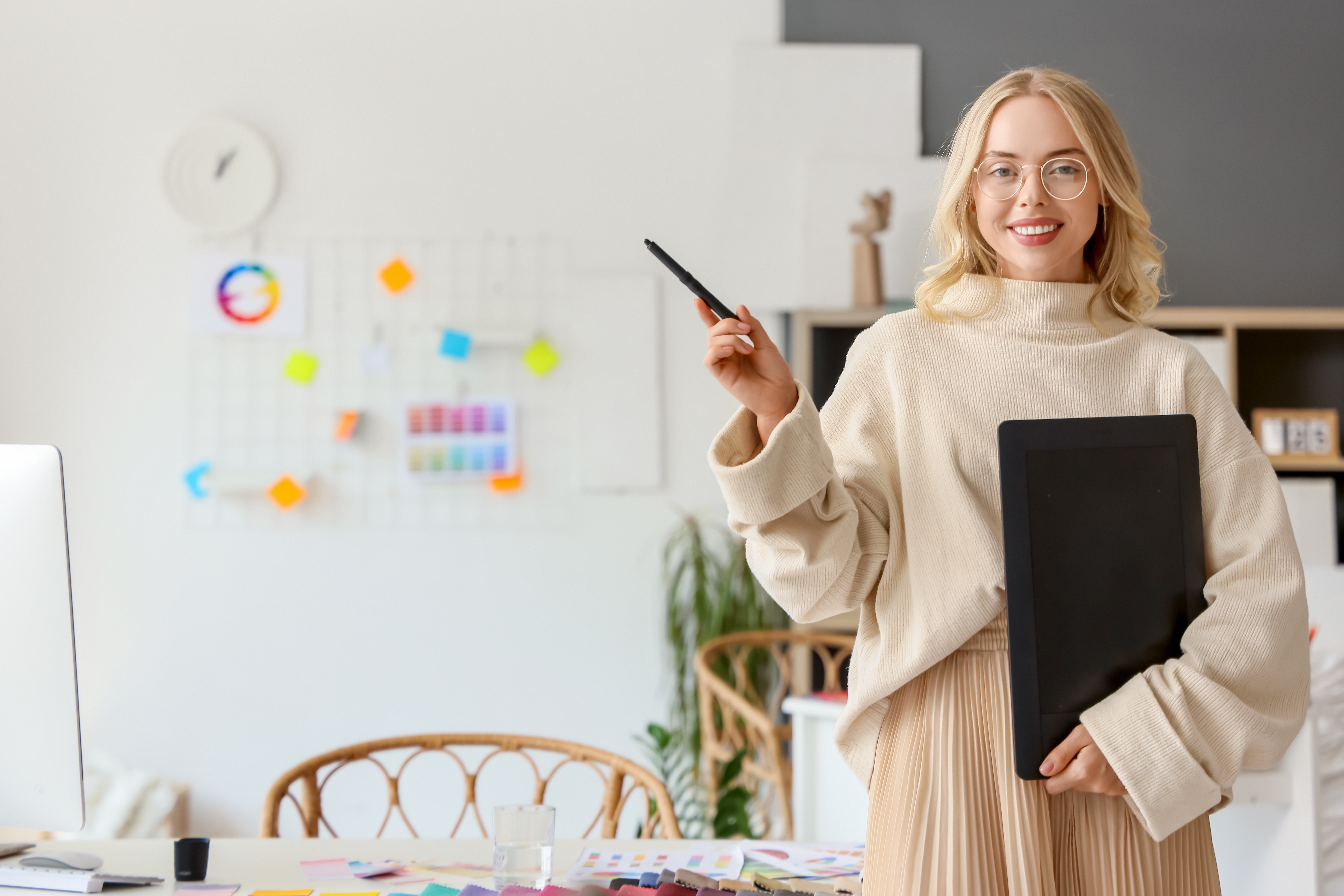 Eine lächelnde Frau mit Brille trägt einen beigen Pullover und hält einen Ordner in der Hand. Sie steht in einem kreativen Arbeitsumfeld mit einem Schreibtisch und bunten Notizen im Hintergrund.