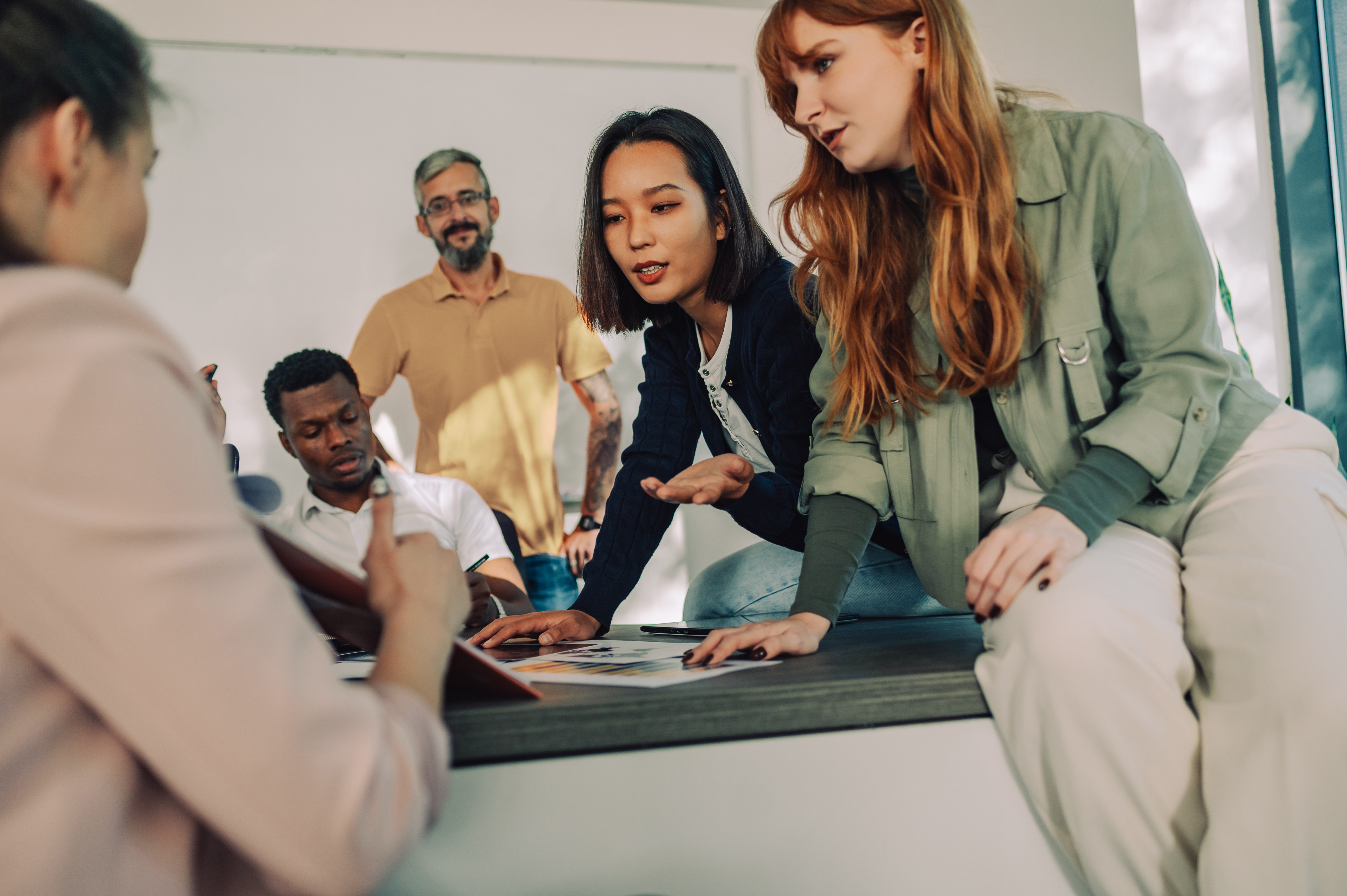 A group of five people is discussing in a modern room. Two women are pointing at documents on a table while a man in the background observes. The atmosphere feels collaborative and engaged.