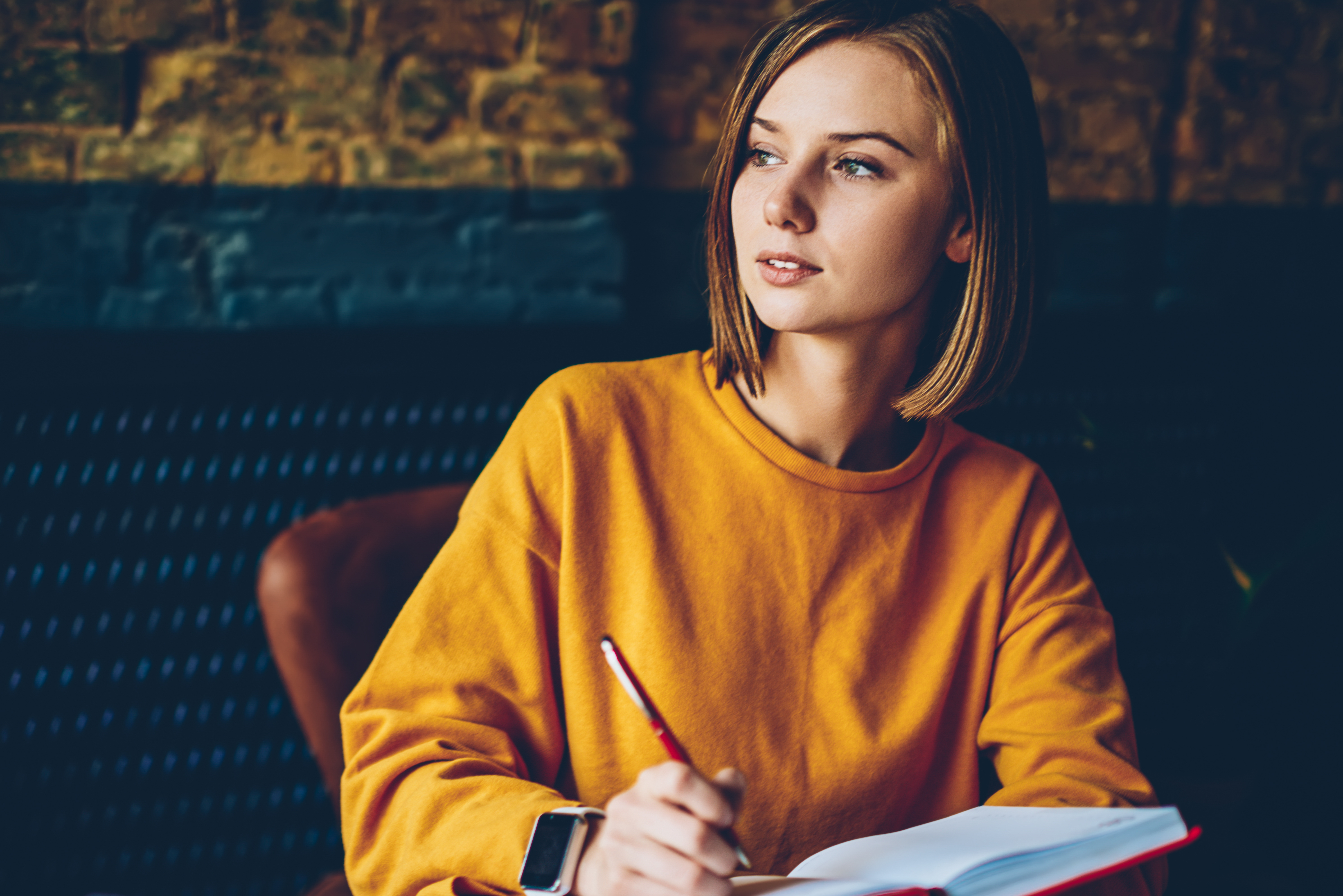Eine junge Frau in einem orangefarbenen Pullover sitzt an einem Tisch, hält einen Stift und schreibt in ein Notizbuch.