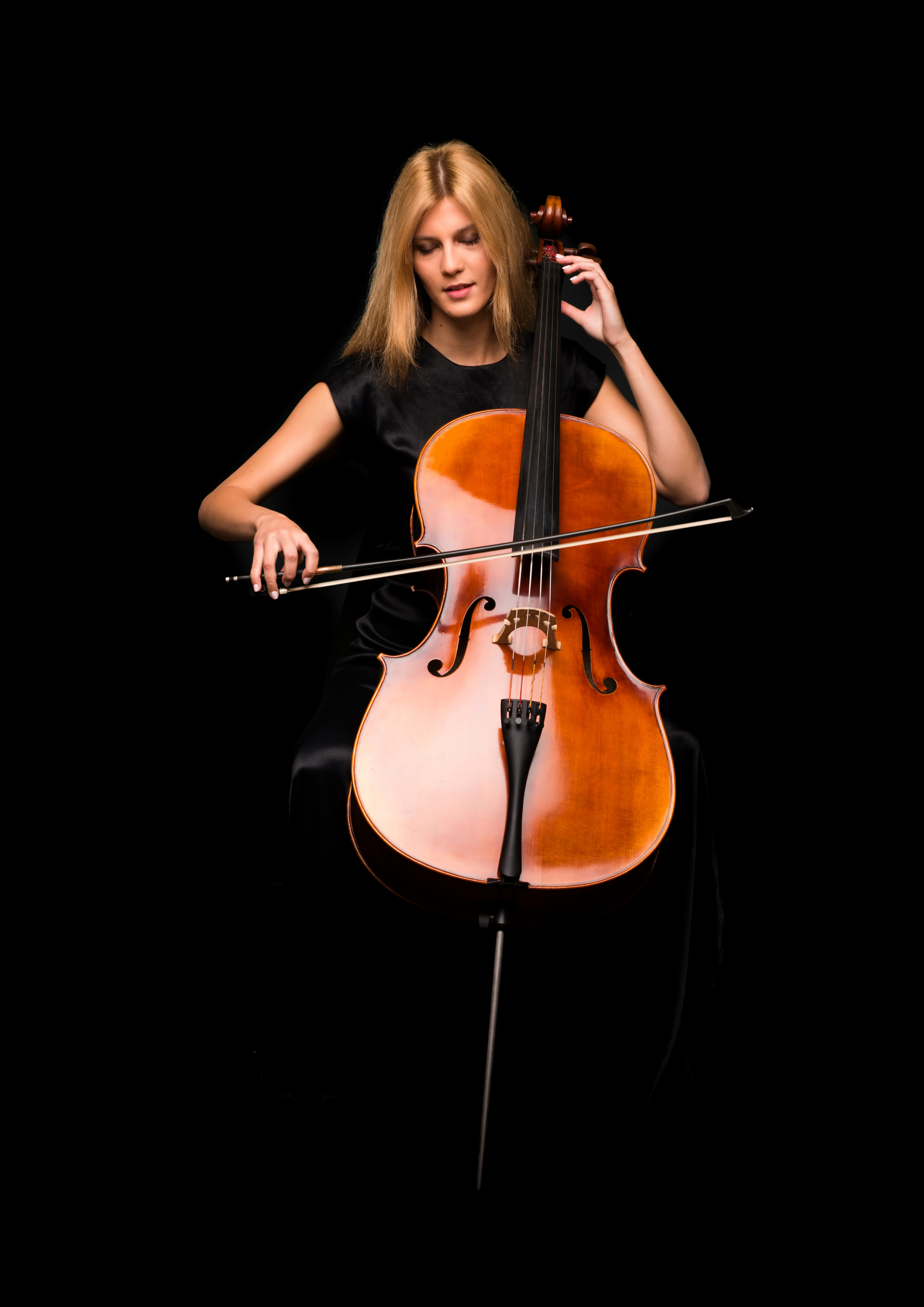 Eine Frau mit langen blonden Haaren spielt ein Cello. Sie sitzt in einem dunklen Raum und hält den Bogen in der rechten Hand, während sie mit der linken Hand die Saiten greift.