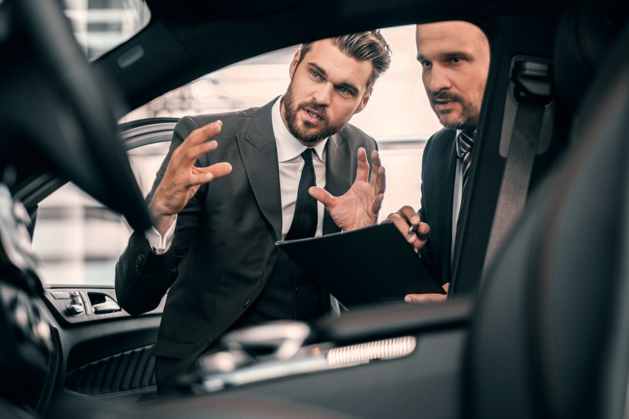 Zwei Männer in Anzügen diskutieren im Innenraum eines Autos. Einer zeigt mit den Händen auf etwas, während der andere Notizen auf einem Tablet macht. Beide scheinen an einem geschäftlichen Gespräch beteiligt zu sein.