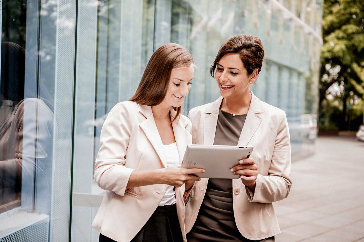 Zwei Frauen in Business-Anzügen stehen draußen und betrachten gemeinsam ein Tablet. Sie lächeln und scheinen sich über den Inhalt des Tablets auszutauschen. Im Hintergrund sind moderne Gebäude und Bäume sichtbar.