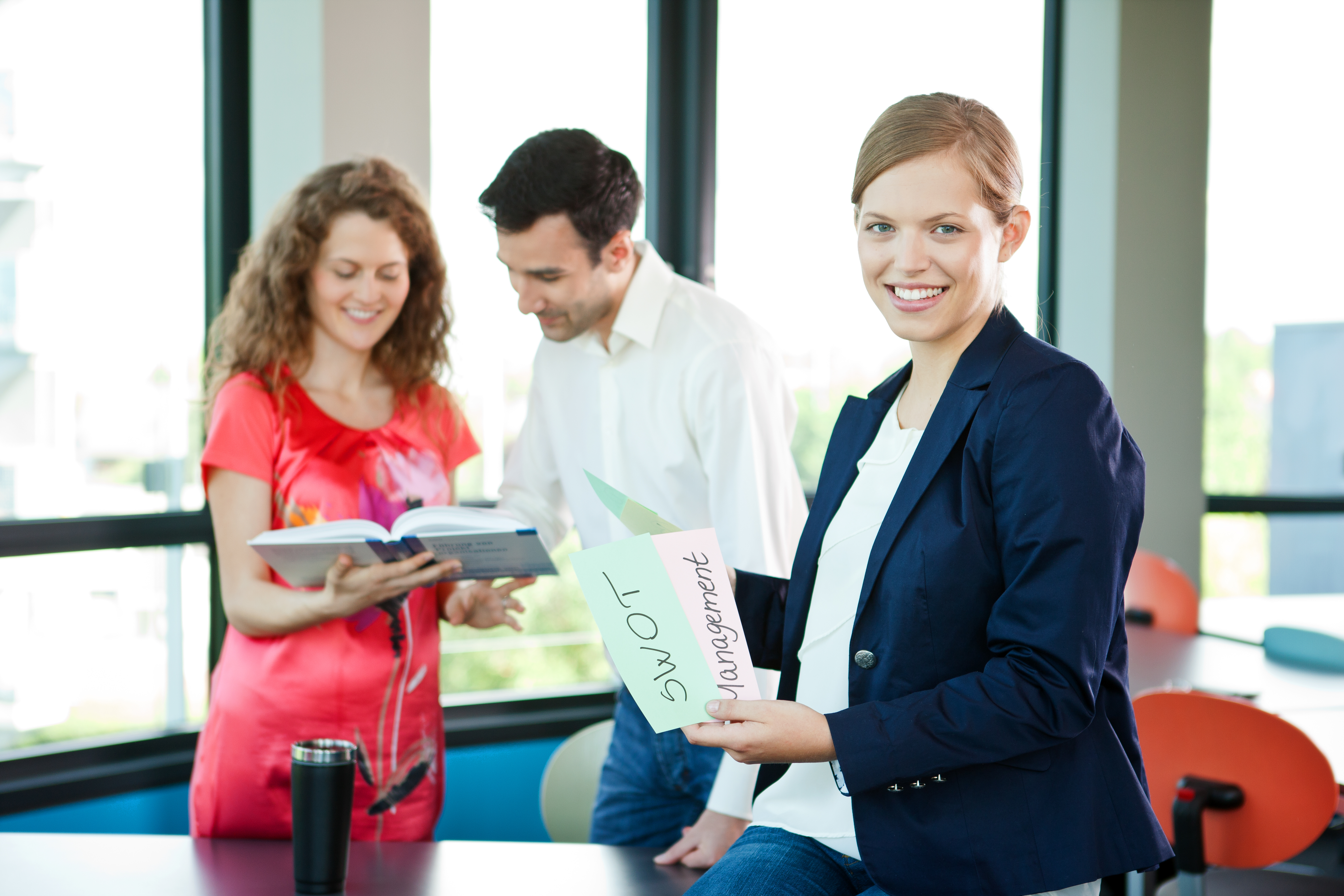 Drei Personen in einem modernen Büro, eine Frau hält ein Blatt mit der Aufschrift "SWOT Management", während die anderen beiden ein Buch betrachten.