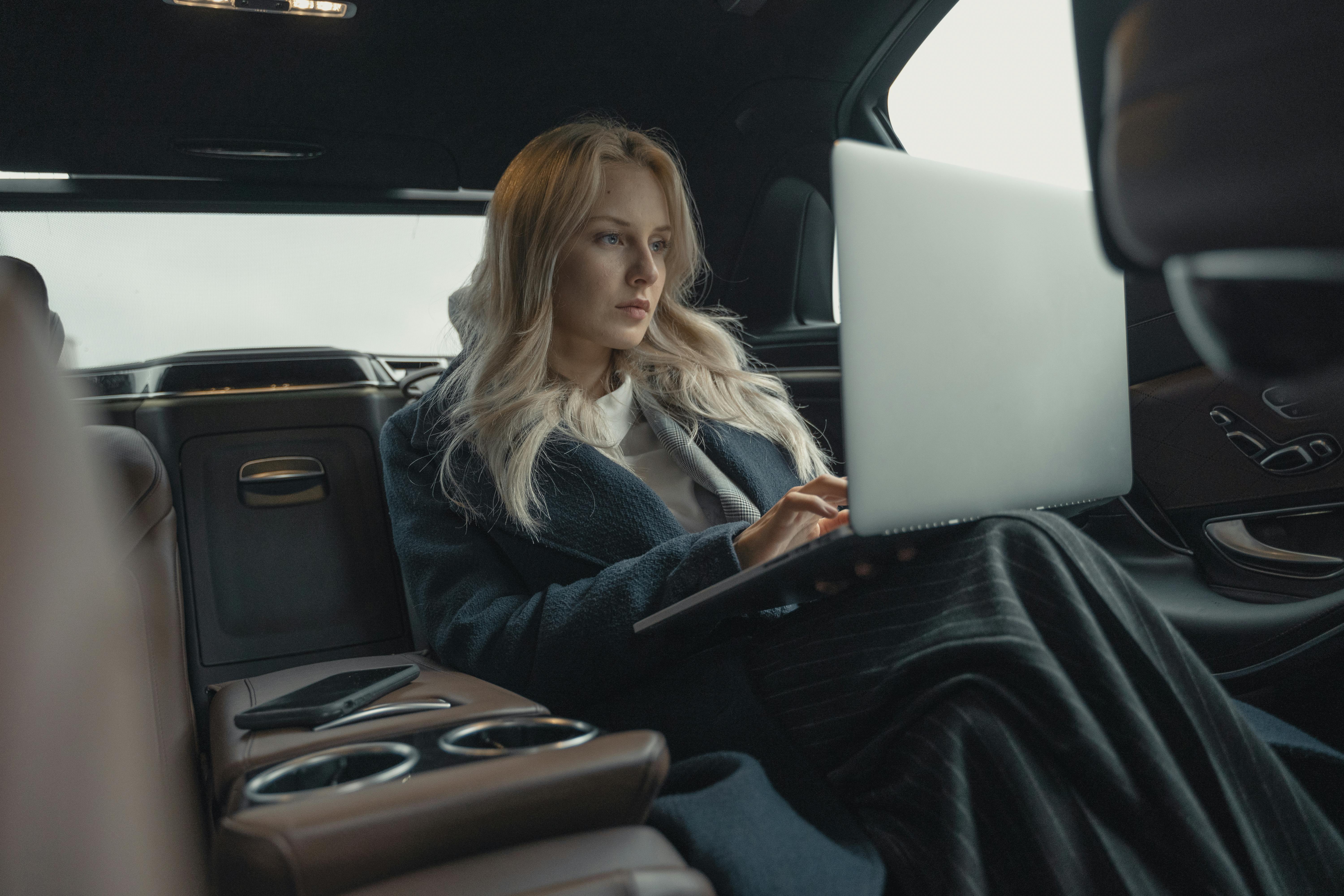 Eine Frau mit langen blonden Haaren sitzt in einem luxuriösen Auto und arbeitet an einem Laptop. Sie trägt einen langen, dunklen Mantel und ist auf den Bildschirm konzentriert. Neben ihr liegt ein Smartphone.