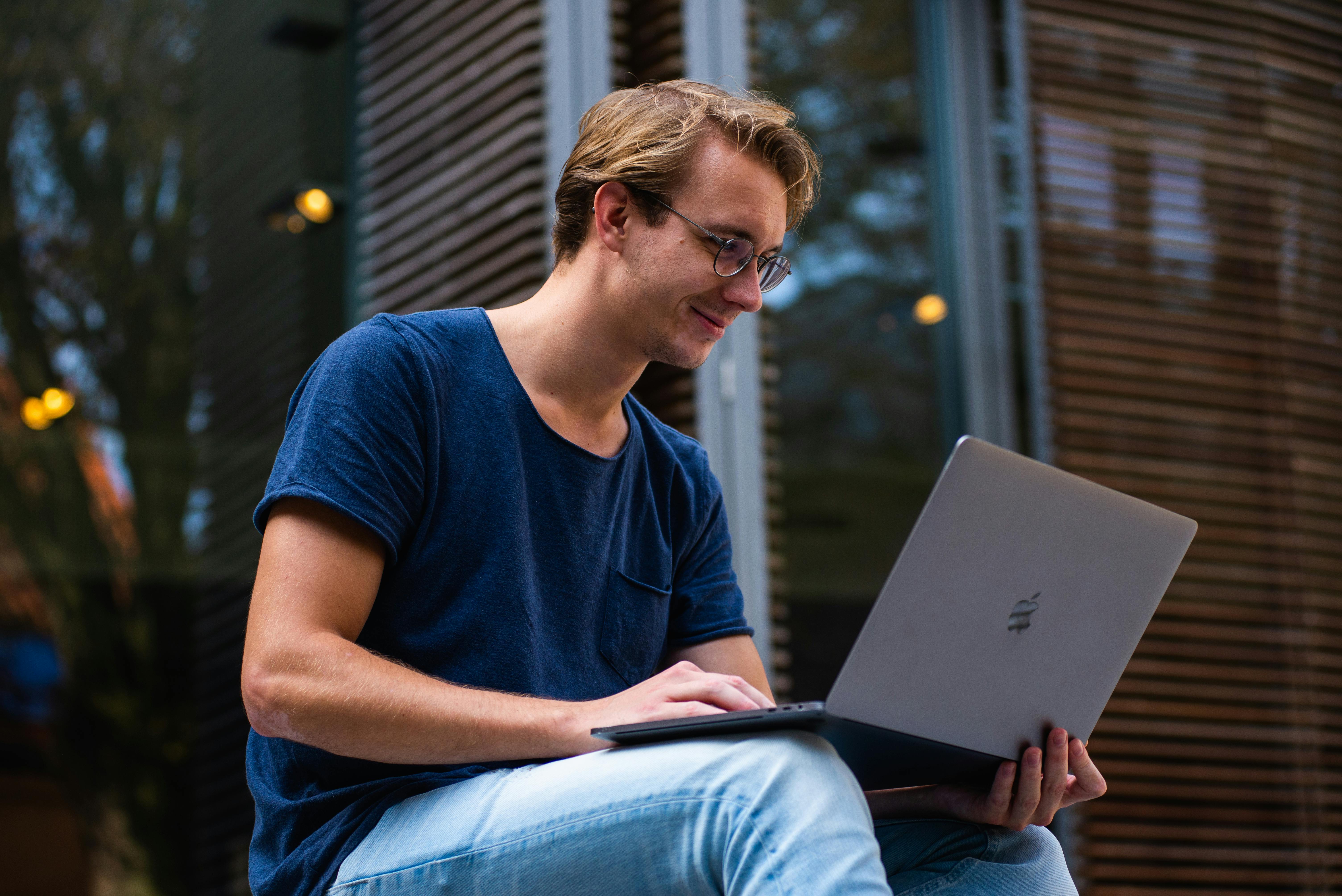 A young man is sitting outside, working on a laptop. He is wearing a blue T-shirt and glasses. The background features wooden slats and blurred trees.