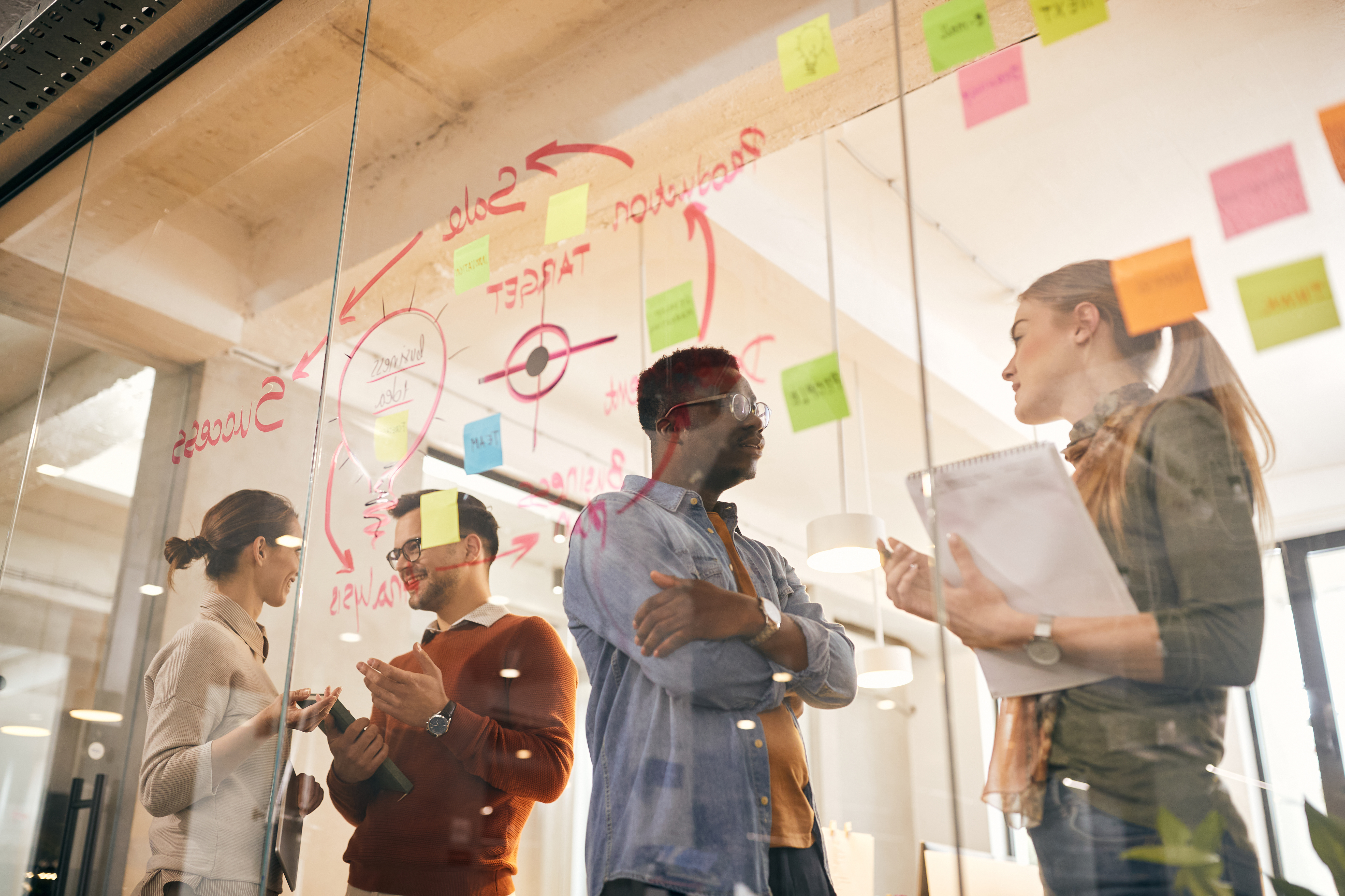 A team of four people is discussing in front of a glass wall covered with colorful sticky notes and diagrams. Two of them are taking notes while one person is speaking, and the fourth person is listening attentively.