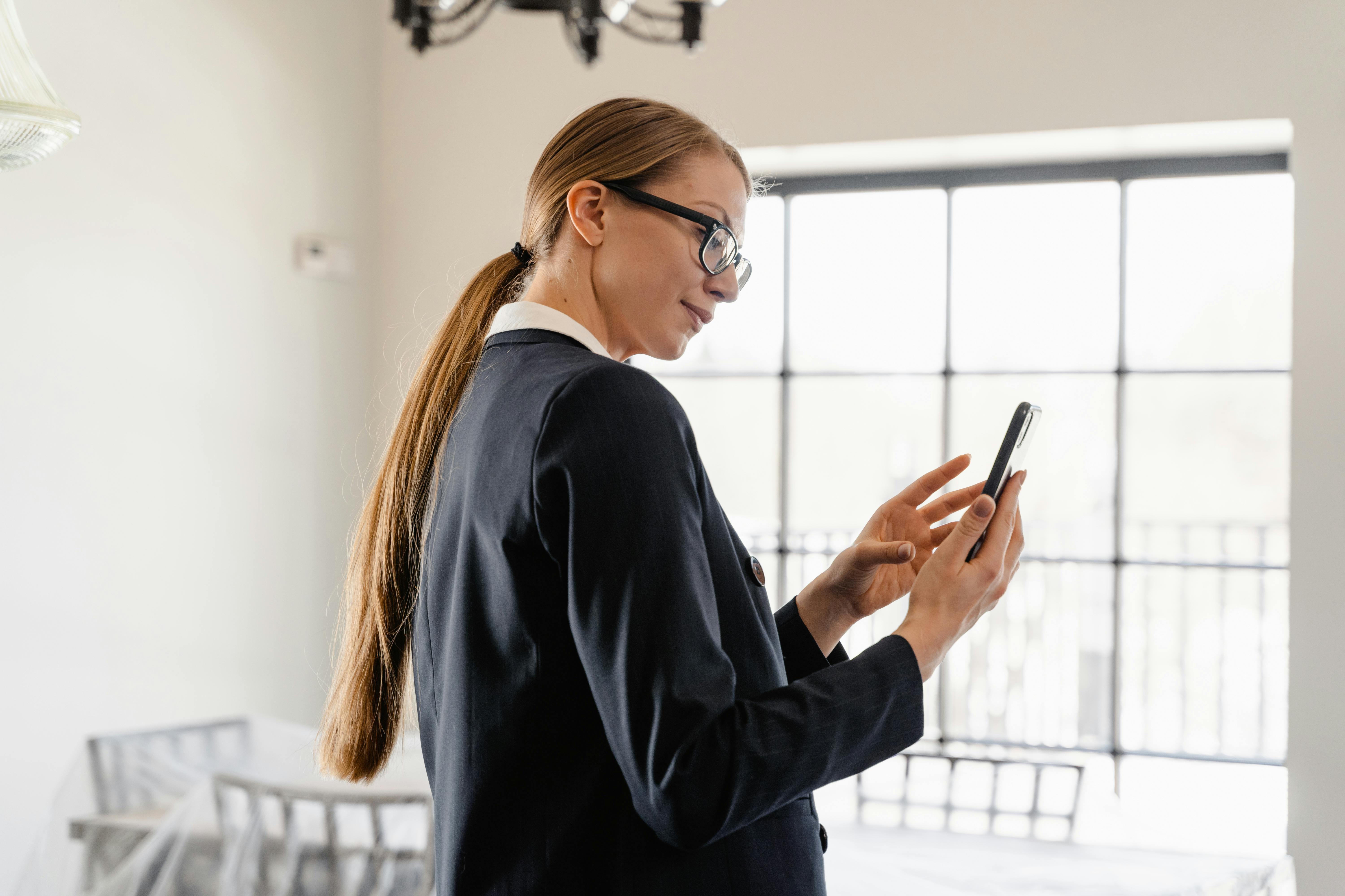 Eine Frau mit langen, glatten Haaren und Brille trägt einen Anzug und schaut auf ihr Smartphone. Sie steht in einem hellen Raum mit großen Fenstern und einem modernen, minimalistischen Design.