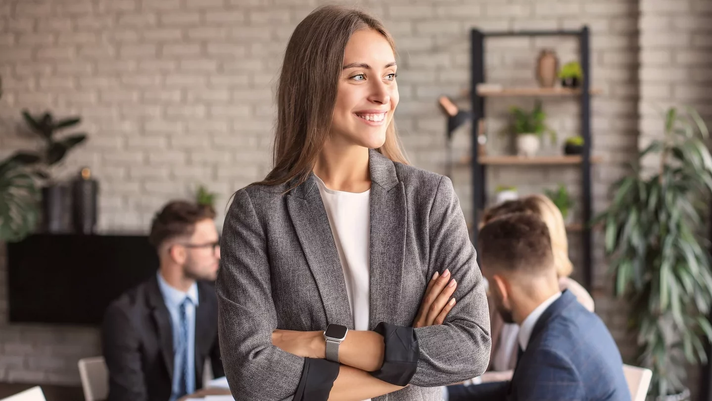 A smiling woman in a gray blazer stands with her arms crossed in a modern office. In the background, several people in formal attire can be seen sitting at a table and conversing with each other. Plants decorate the space.
