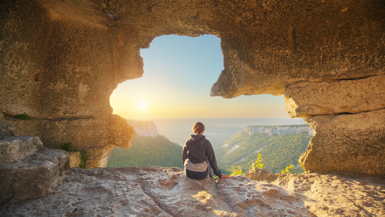 A person is sitting on a rock in a cave, gazing at a landscape with mountains and a sunset. The scene features a natural opening in the rock wall that reveals the surroundings.