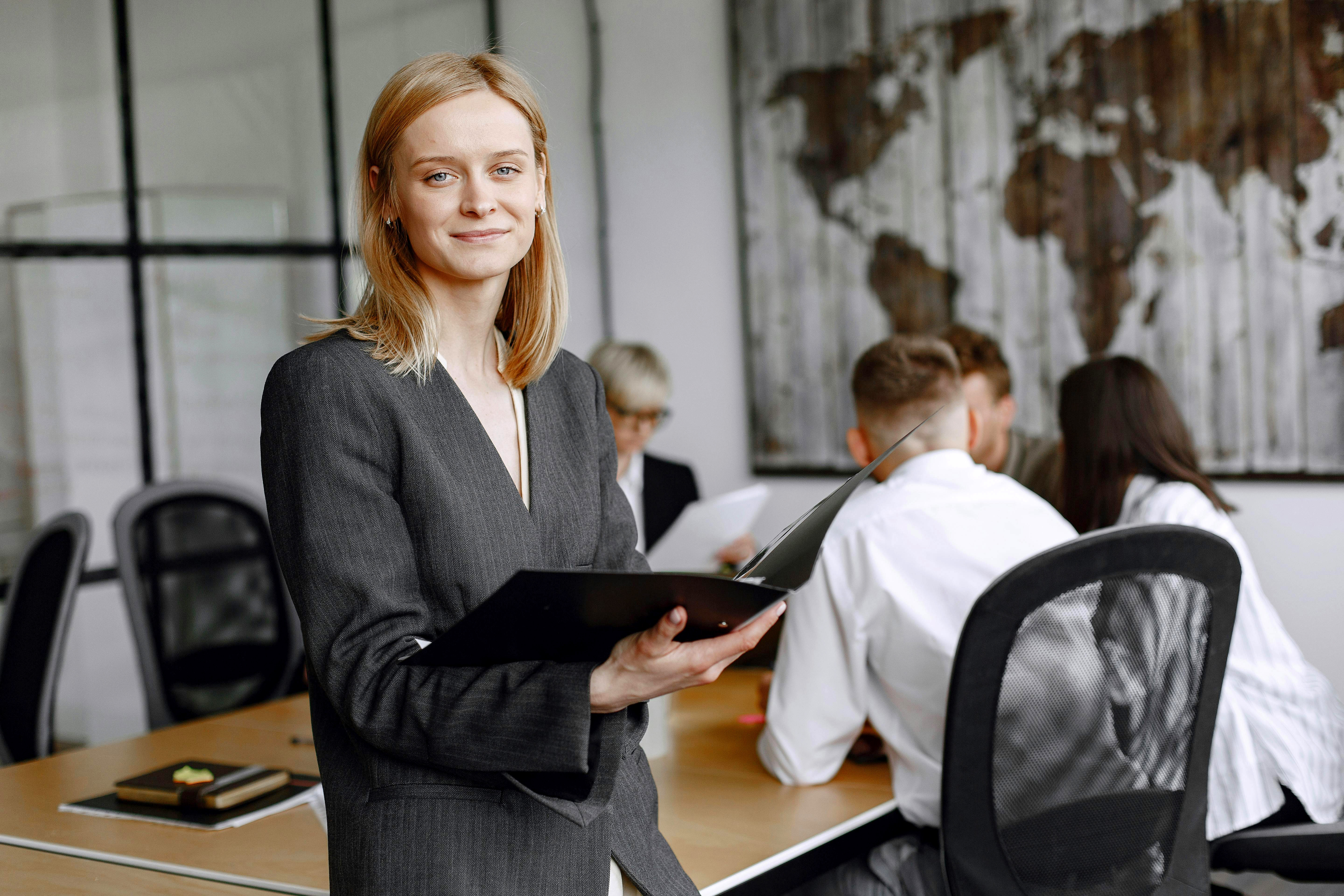 Eine Frau in einem grauen Anzug hält ein Notizbuch und lächelt, während im Hintergrund eine Gruppe von Personen in einem Meeting sitzt.