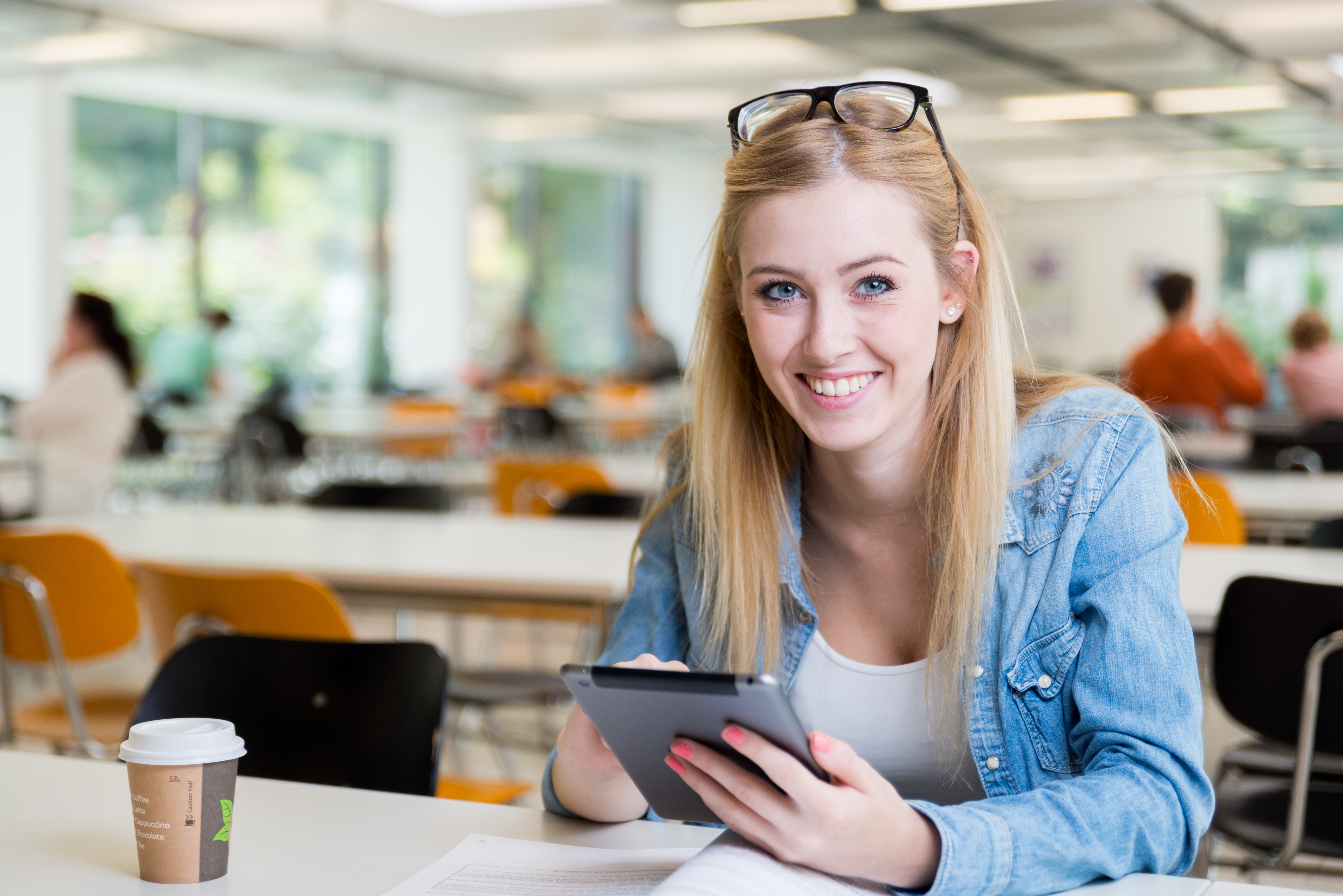 Eine blonde Frau mit Brille sitzt an einem Tisch, lächelt und nutzt ein Tablet. Eine Kaffeetasse steht neben ihr.