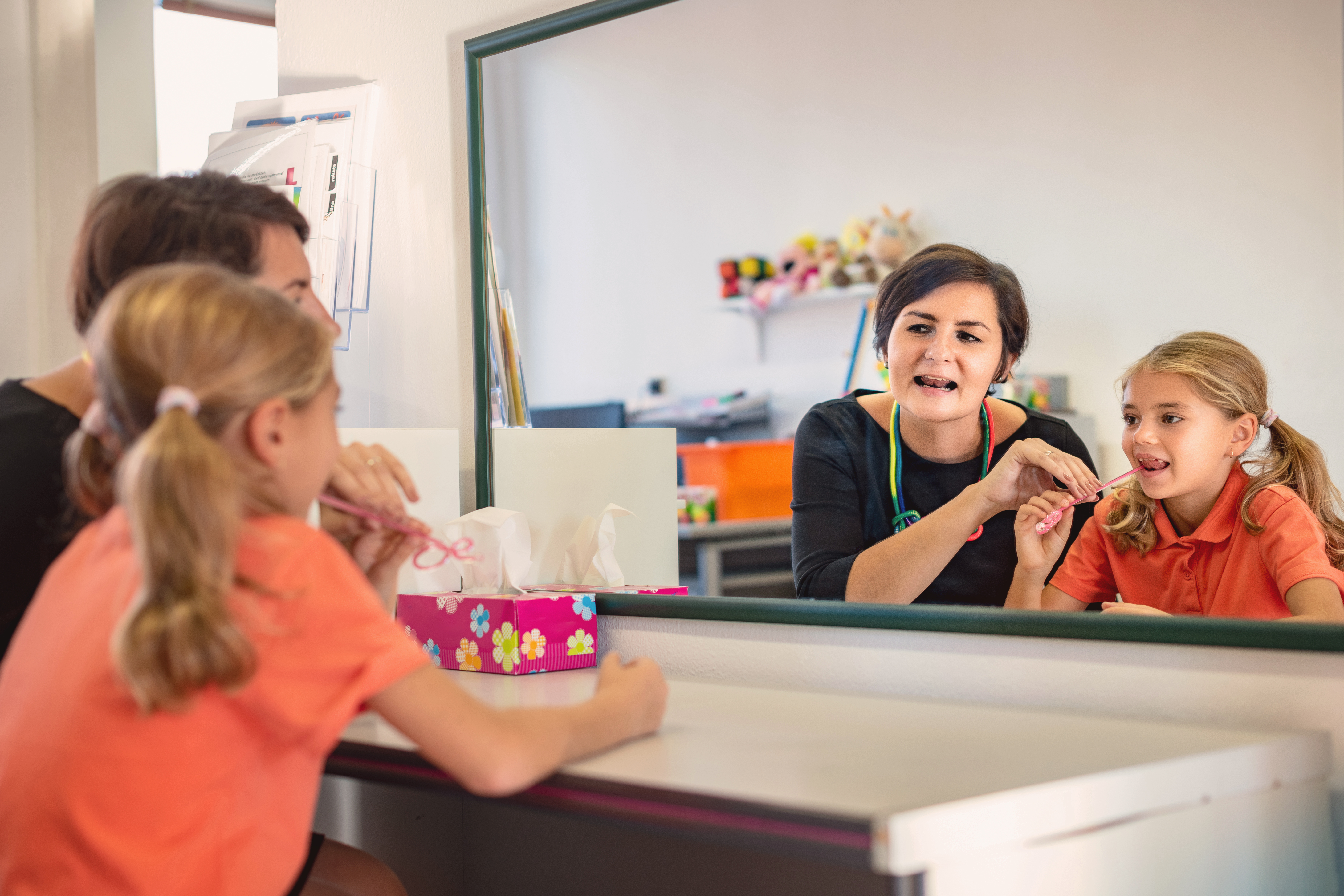 A woman is explaining something to a girl in an orange shirt in front of a mirror. The girl is holding a toothbrush in her hand. On the table, there is a box of tissues, and in the background, there are some toys.