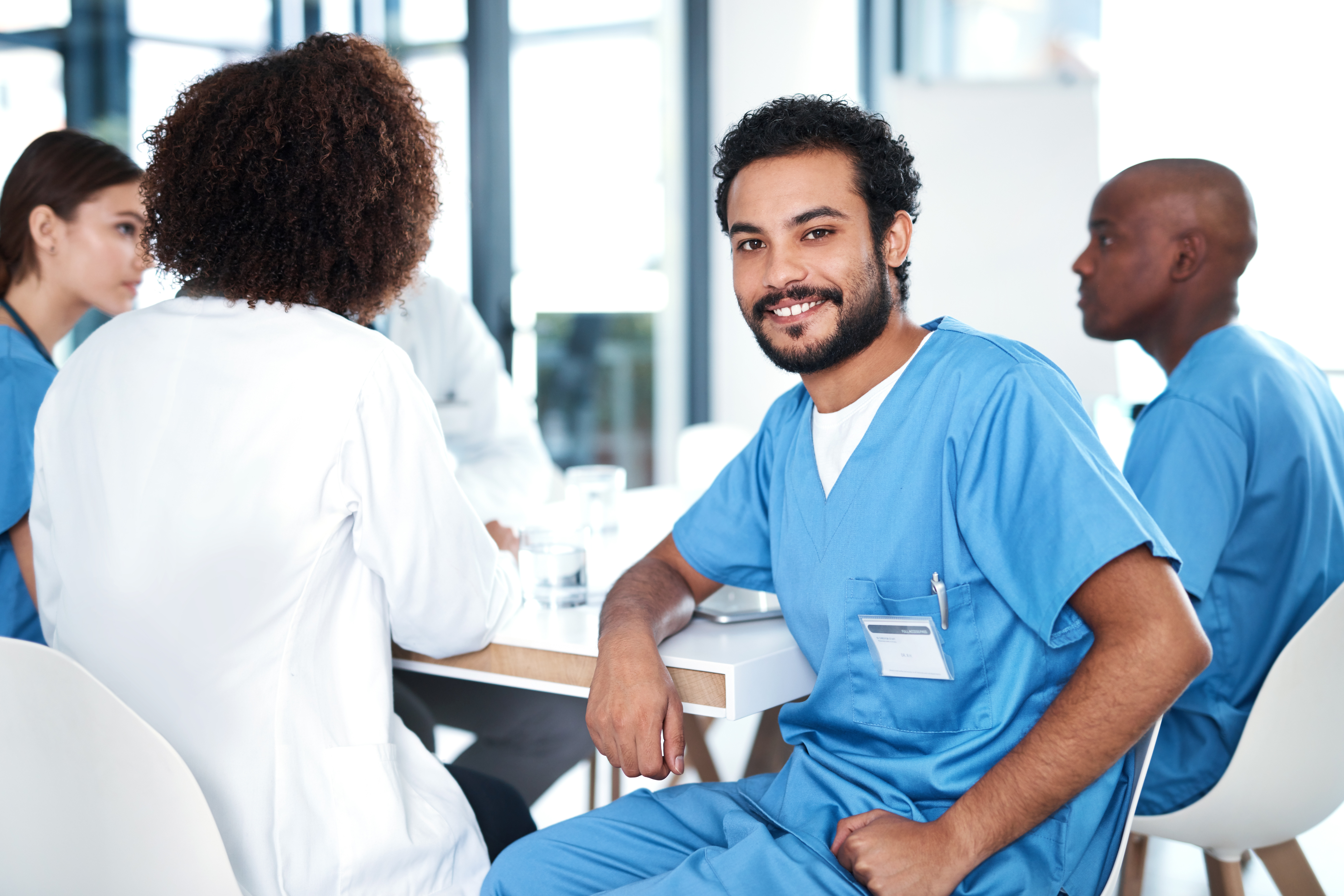 A young man in blue medical attire is sitting at a table, smiling. Around him are several people in similar clothing participating in a meeting. The room has large windows that let in plenty of natural light.