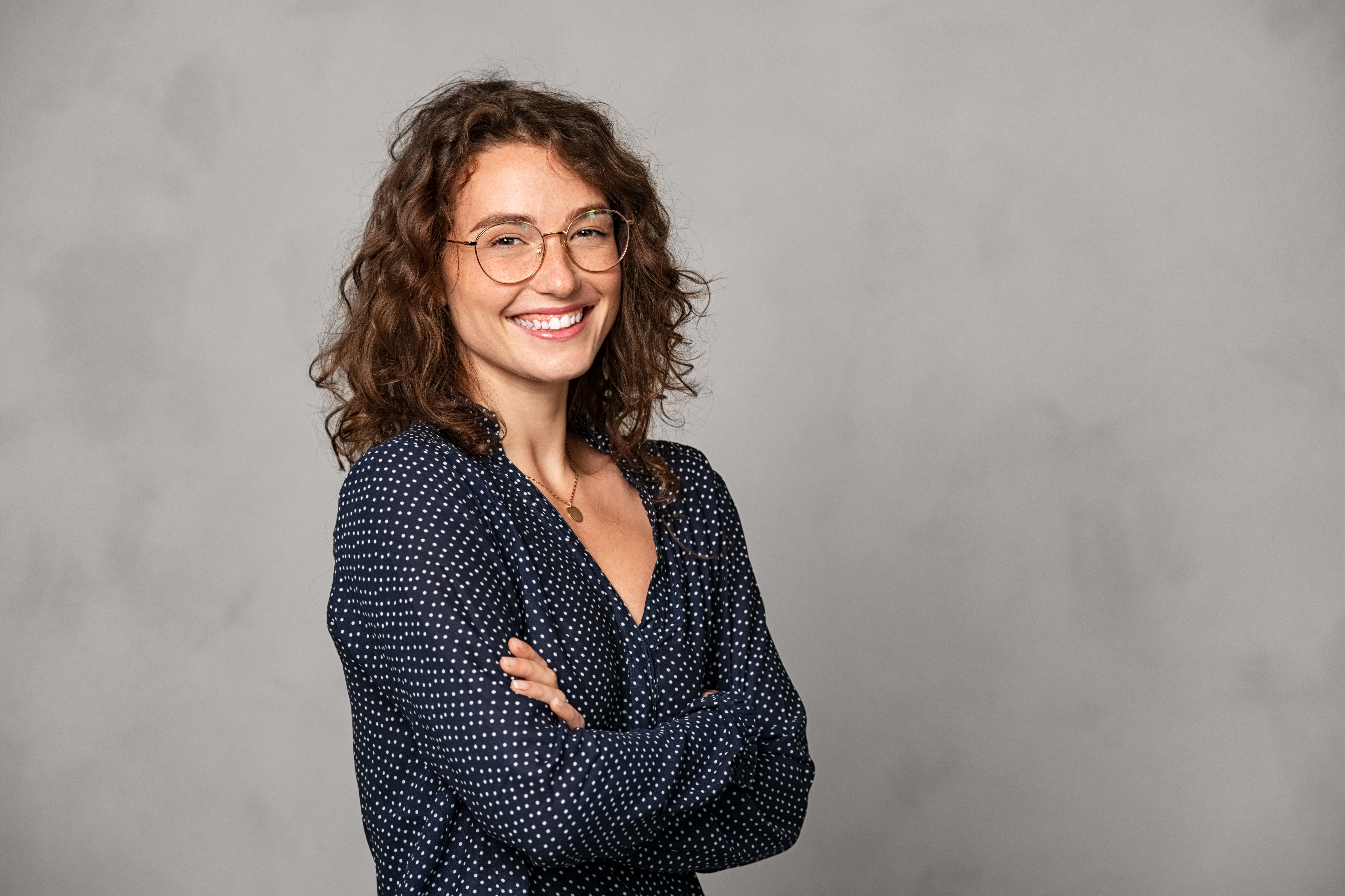 A smiling woman with curly brown hair is wearing a polka-dotted blouse and a necklace. She stands with her arms crossed in front of a gray background. Her glasses highlight her friendly face.