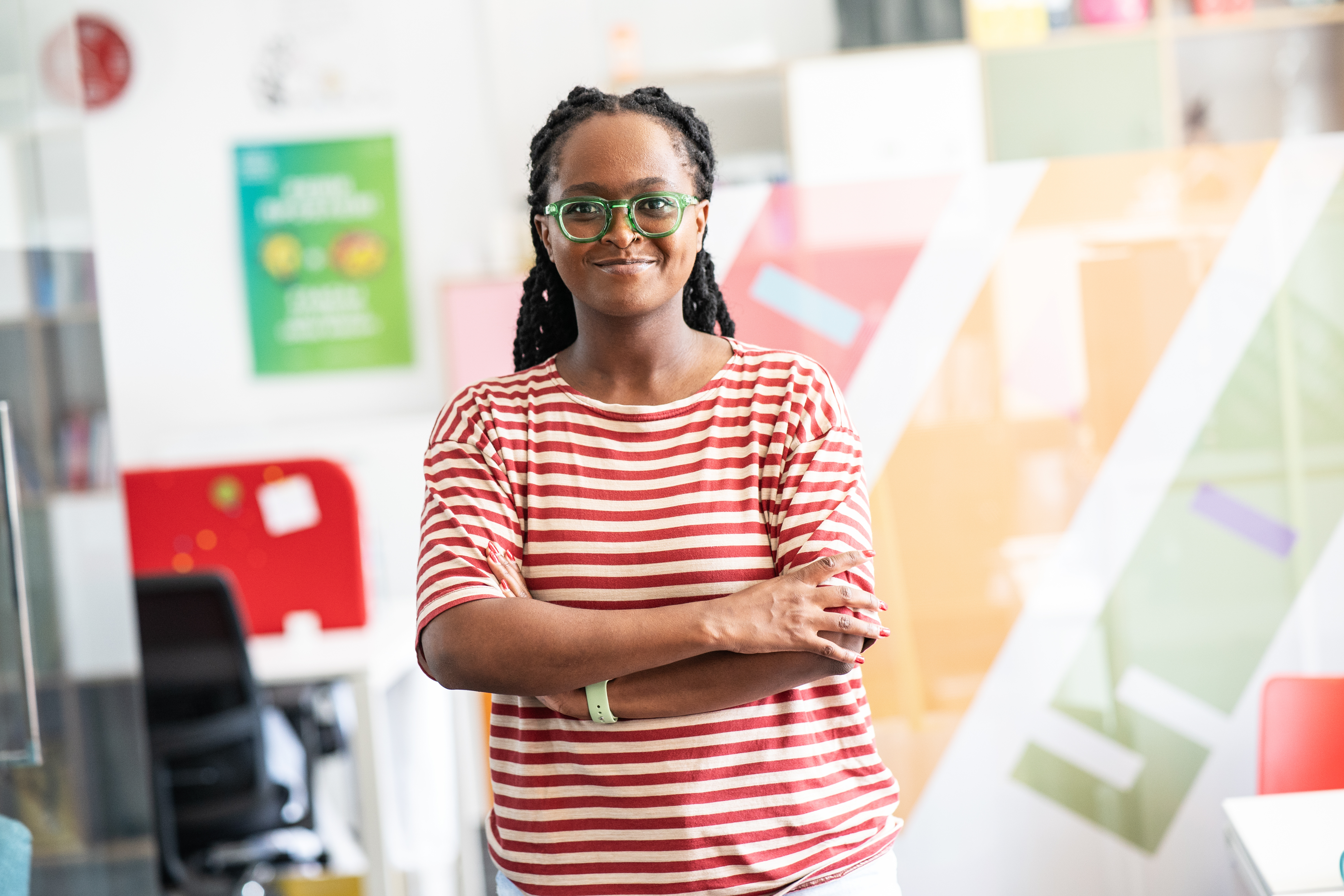 A woman with long, braided hair is wearing a striped T-shirt and standing with her arms crossed in a modern office. She is wearing glasses and has a slight smile, with colorful walls visible in the background.