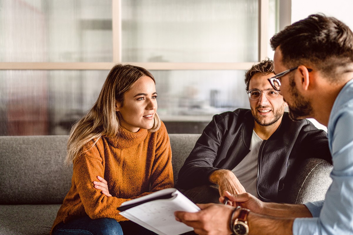 Two people are sitting on a sofa and having a conversation, while a third person is taking notes. The woman is wearing an orange sweater, and the man next to her is in a black jacket. The third man is wearing glasses.