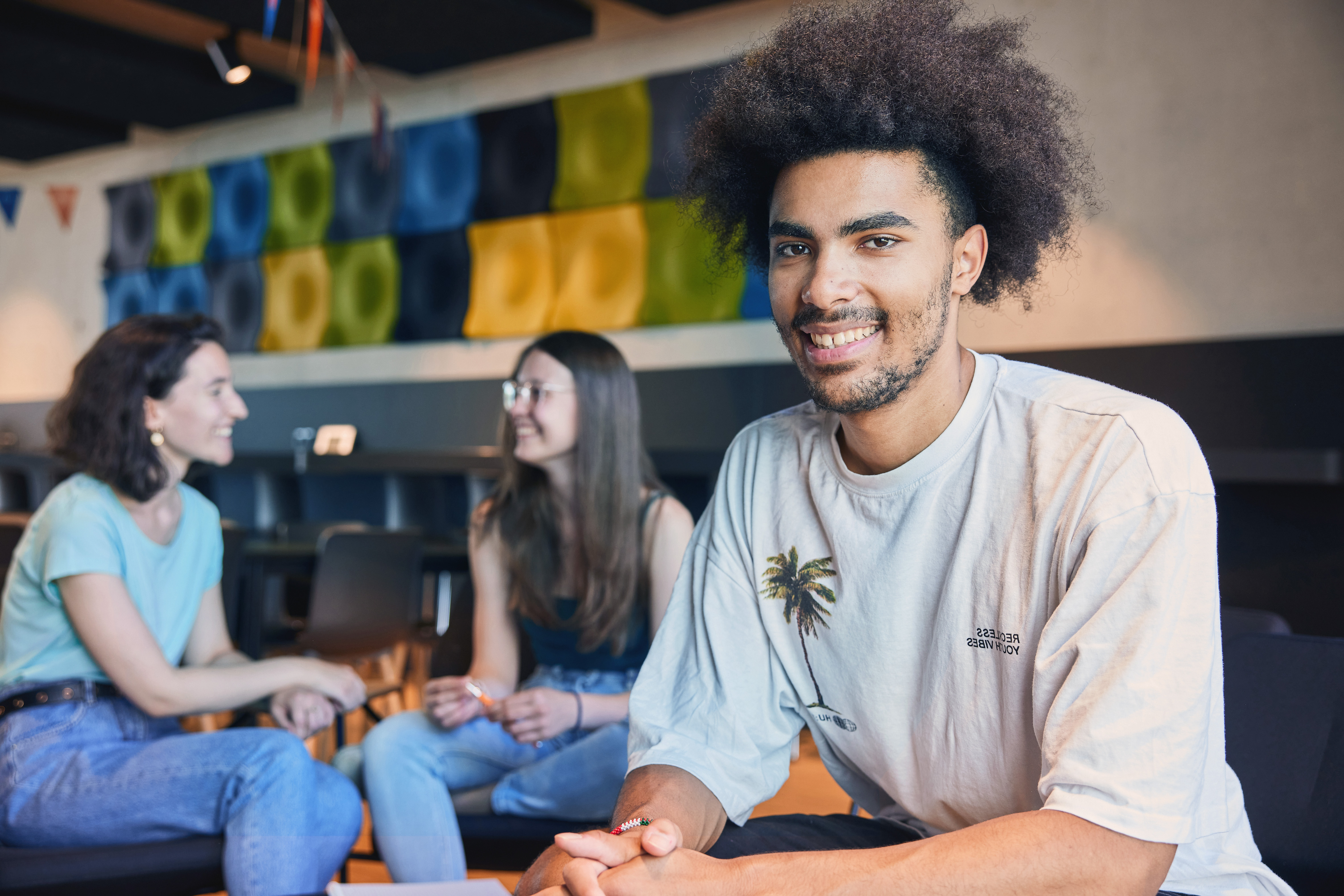 A young man with curly hair sits smiling in the foreground, while two women chat with each other in the background. All three are in a modern space with colorful wall decorations.