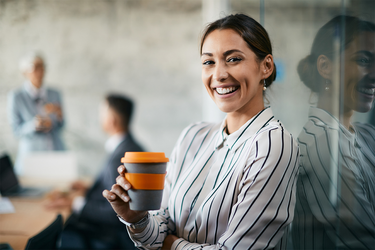 A smiling woman in a striped shirt is holding a coffee cup. In the background, two people can be seen in a meeting room. The atmosphere feels professional and modern.