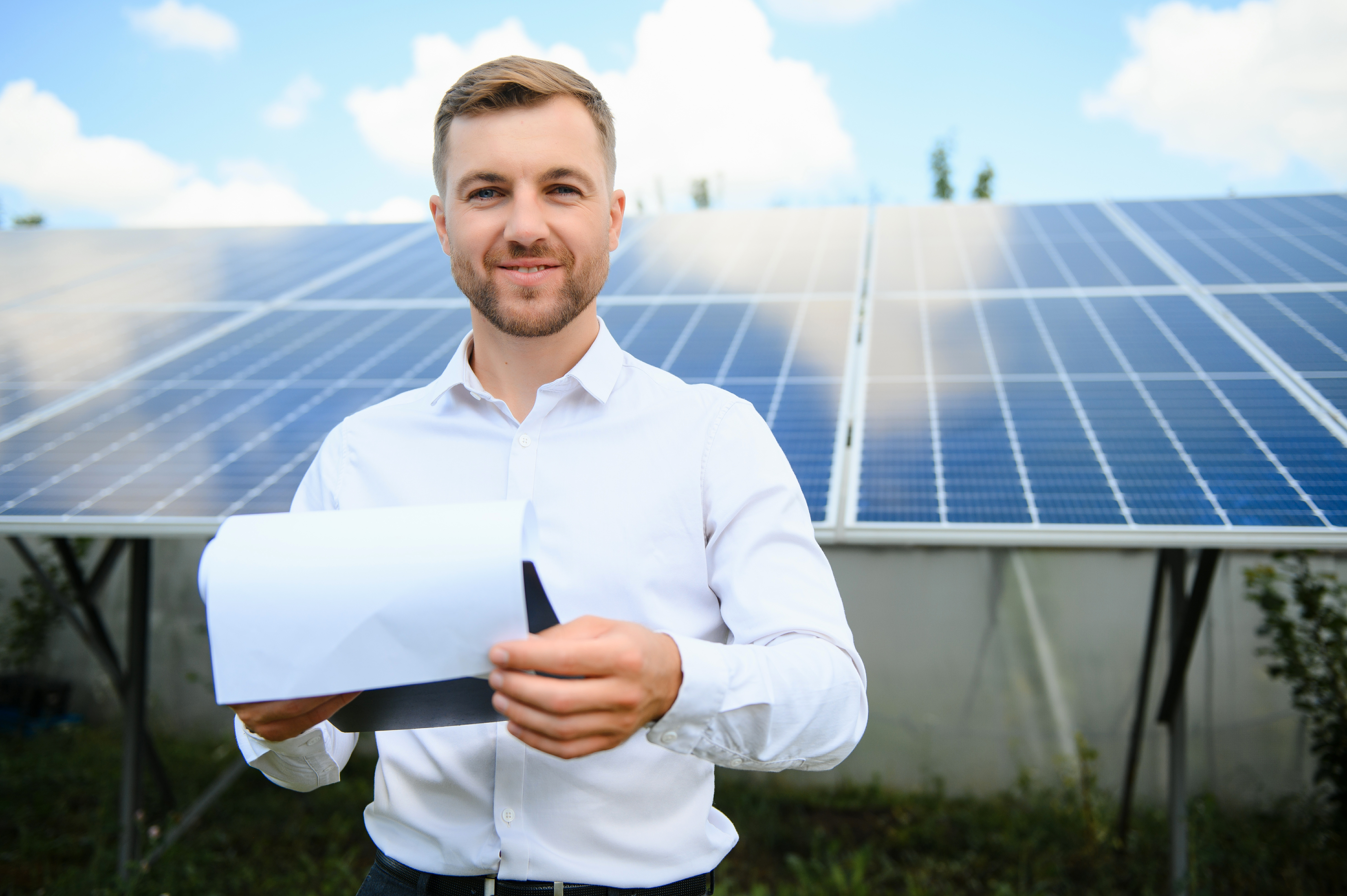 Mann in Hemd hält ein Dokument vor Solarpanelen und lächelt. Die Umgebung zeigt eine sonnige Landschaft mit blauen Himmel.