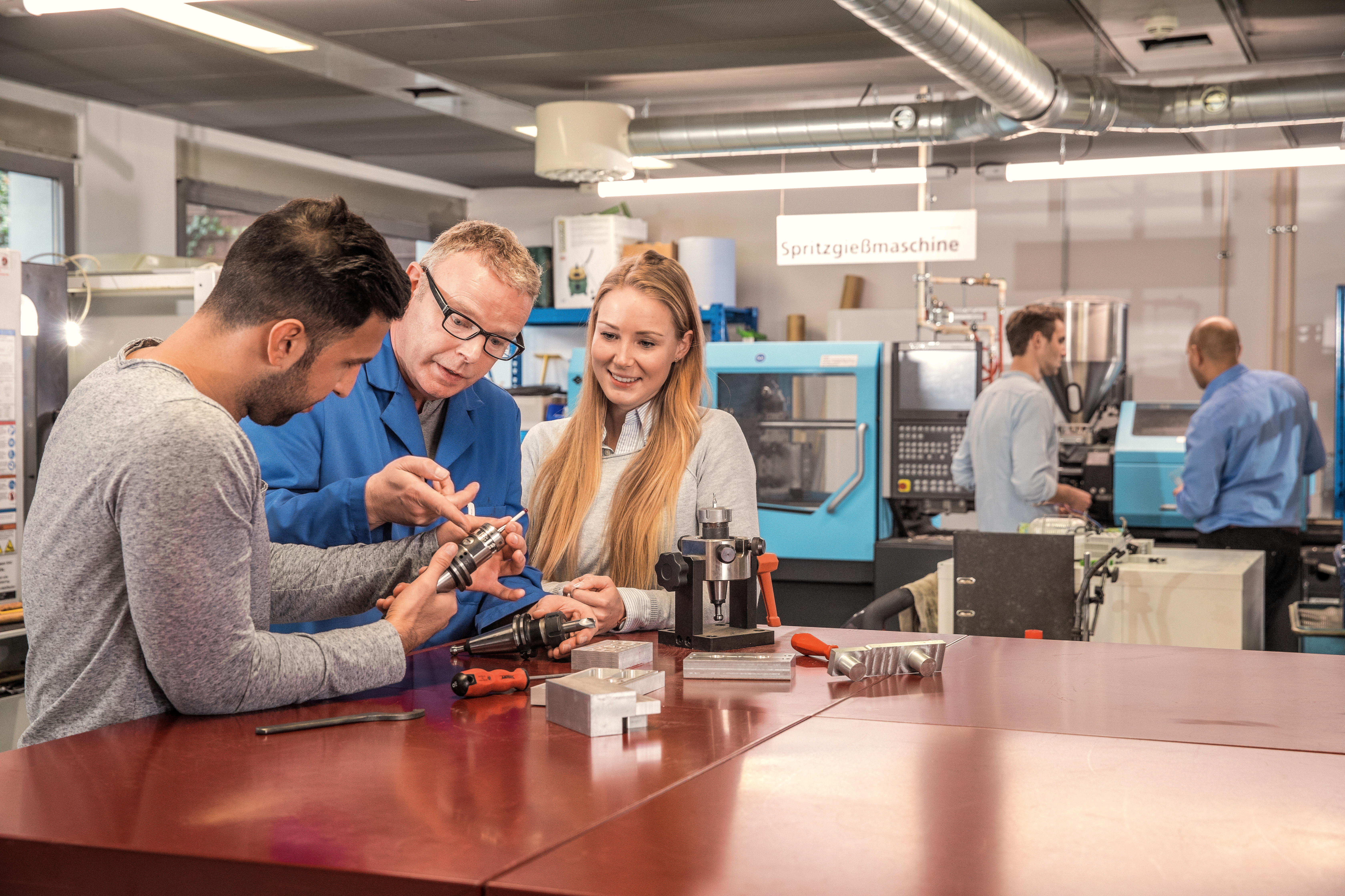 Zwei Personen diskutieren mit einem Lehrer über ein mechanisches Teil an einem Tisch in einer technischen Werkstatt. Im Hintergrund sind Maschinen sichtbar.