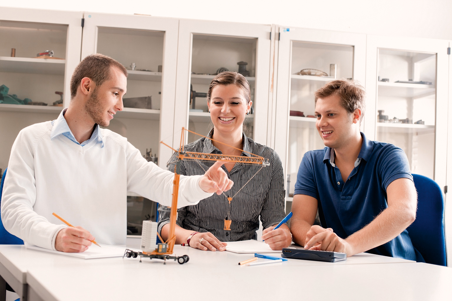 Three people are sitting at a table discussing a model of a crane. One person is pointing at the model while the others listen attentively. Writing materials and a calculator are spread out on the table.
