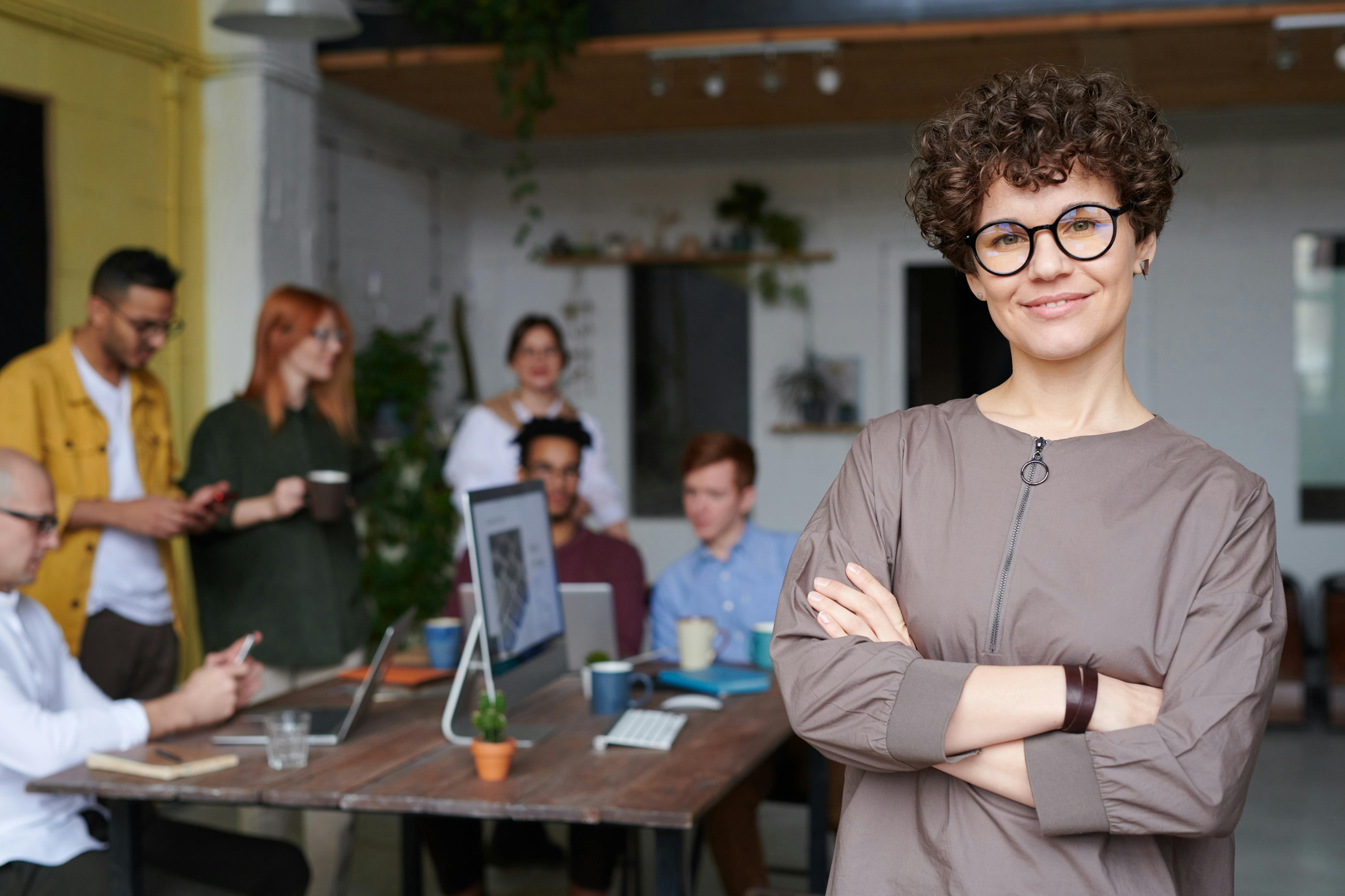 Eine Frau mit lockigem Haar und Brille steht im Vordergrund und lächelt. Im Hintergrund sind mehrere Personen in einem modernen Büro, die an einem Tisch arbeiten und miteinander kommunizieren. Es gibt Laptops und Pflanzen auf dem Tisch.