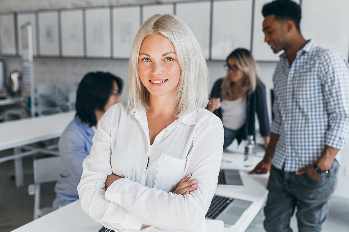 Eine blonde Frau mit schulterlangen Haaren steht in einem modernen Büro und lächelt, während sie die Arme verschränkt. Im Hintergrund sind drei weitere Personen zu sehen, die an Tischen arbeiten und miteinander sprechen.