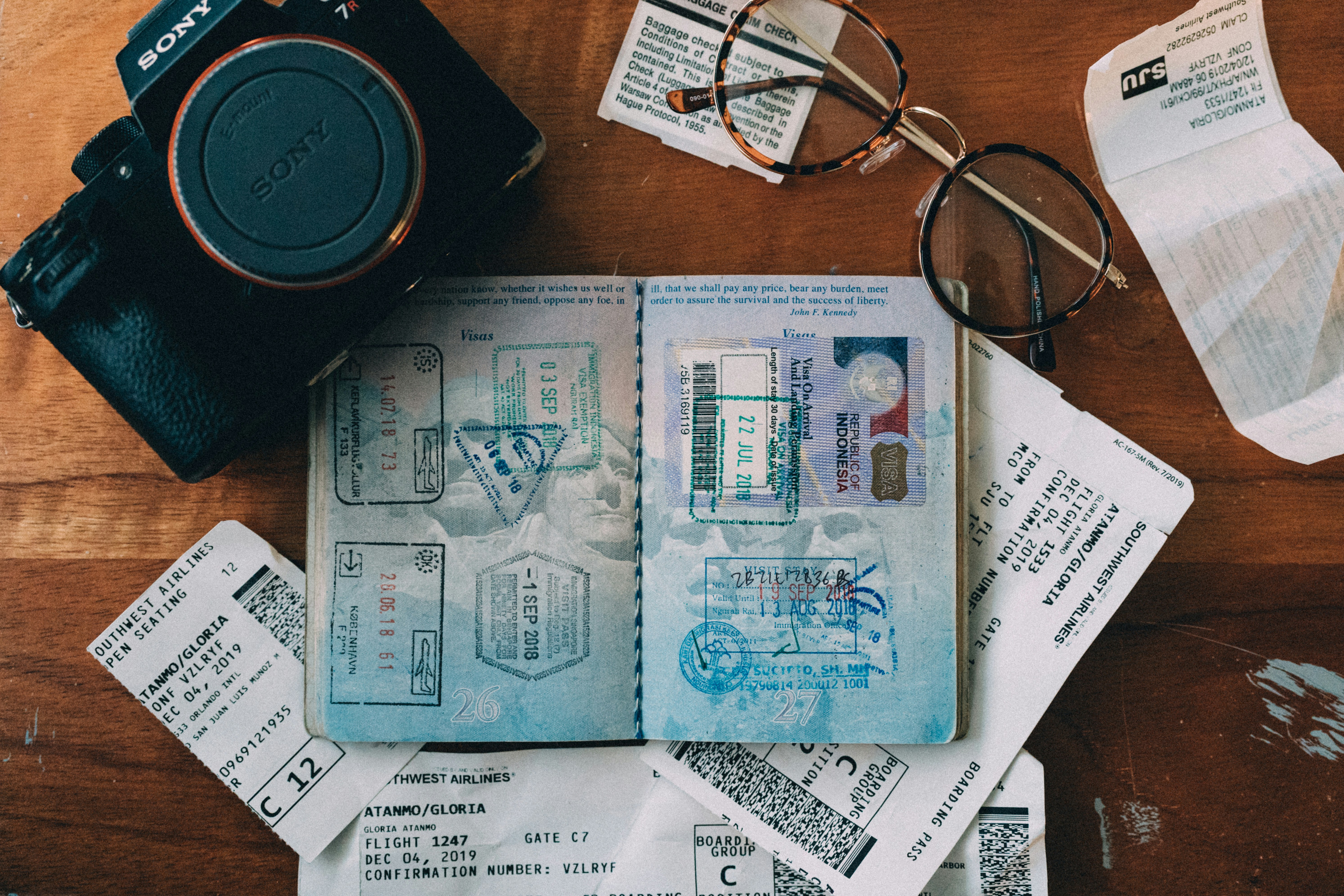 Passport with stamps, flight tickets, and a camera are laid out on a wooden table. A pair of glasses is also visible. The tickets display details about flights and gate information.