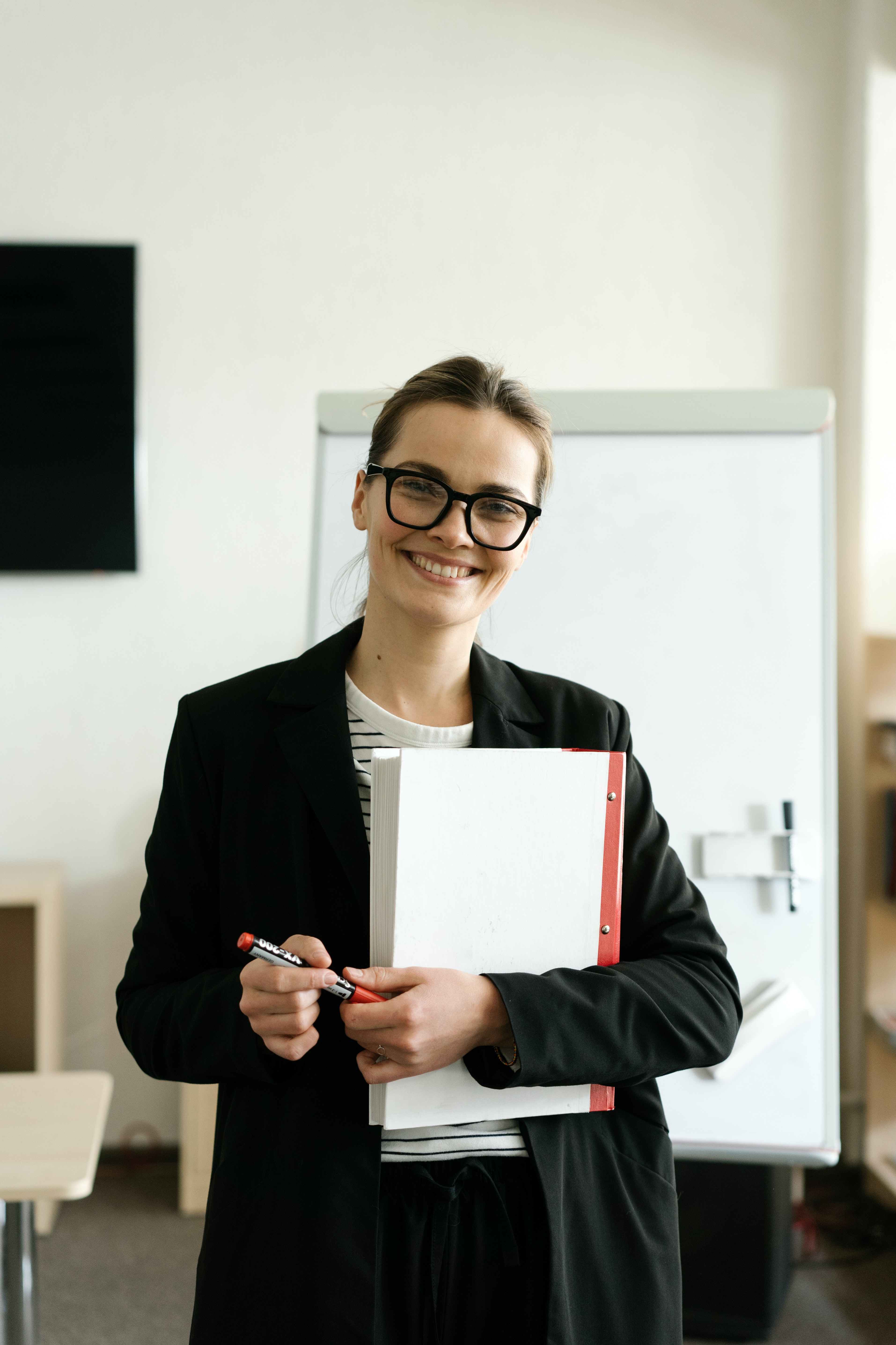 A woman with glasses stands in a room, holding a binder and a marker in her hand. She is wearing a black blazer and a striped top. In the background, a flip chart is visible.